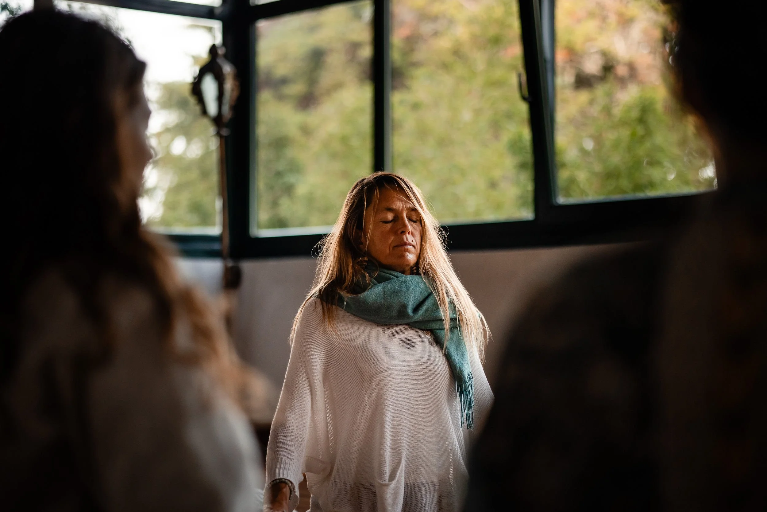 A woman with long blonde hair standing with eyes closed inside a room with large open windows showing trees outside. Two other blurred women are partially visible in the foreground.