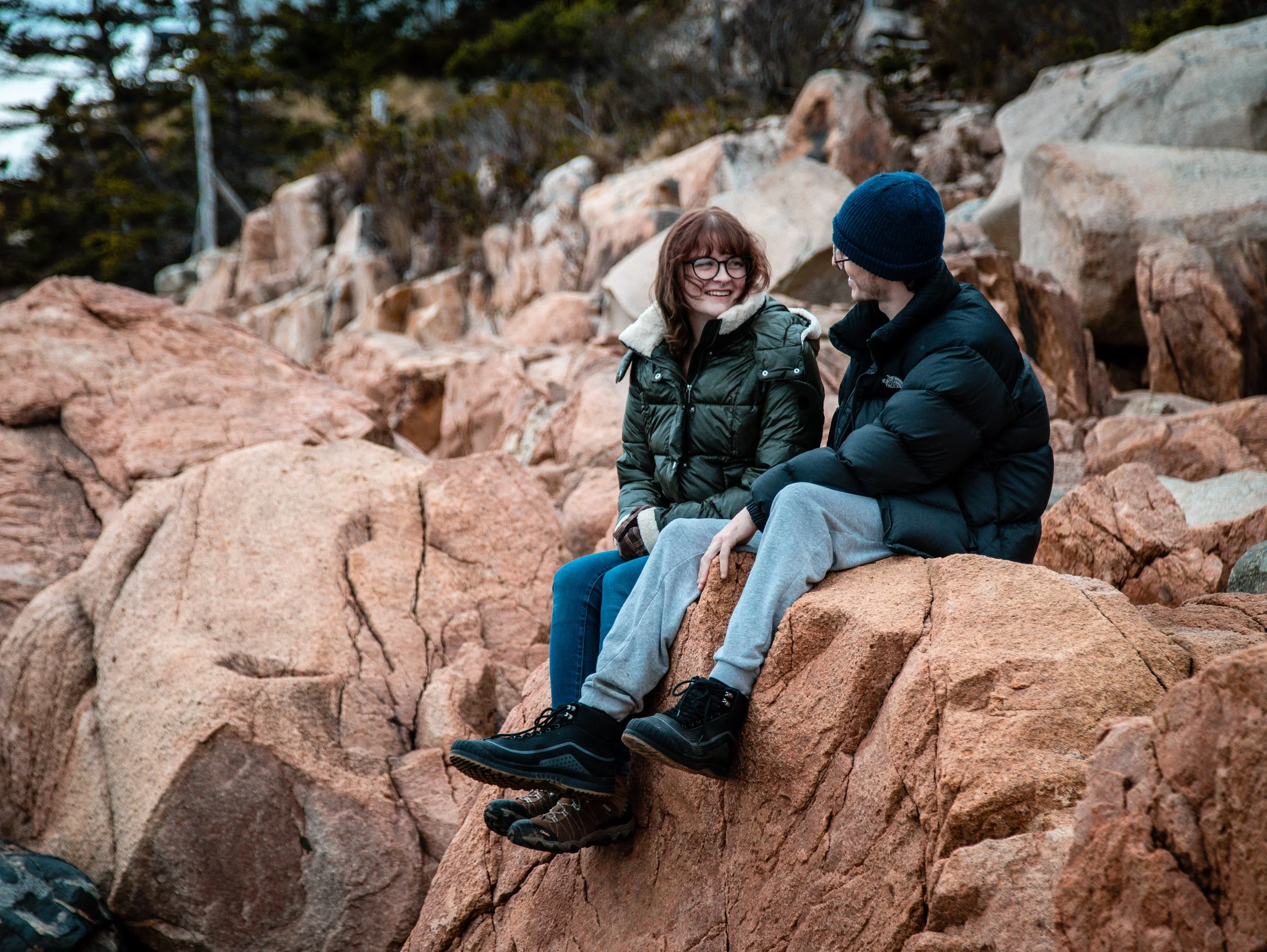 A man and a woman sitting on a rocky landscape, dressed in winter jackets and boots, smiling at each other. The background features large rocks and trees.