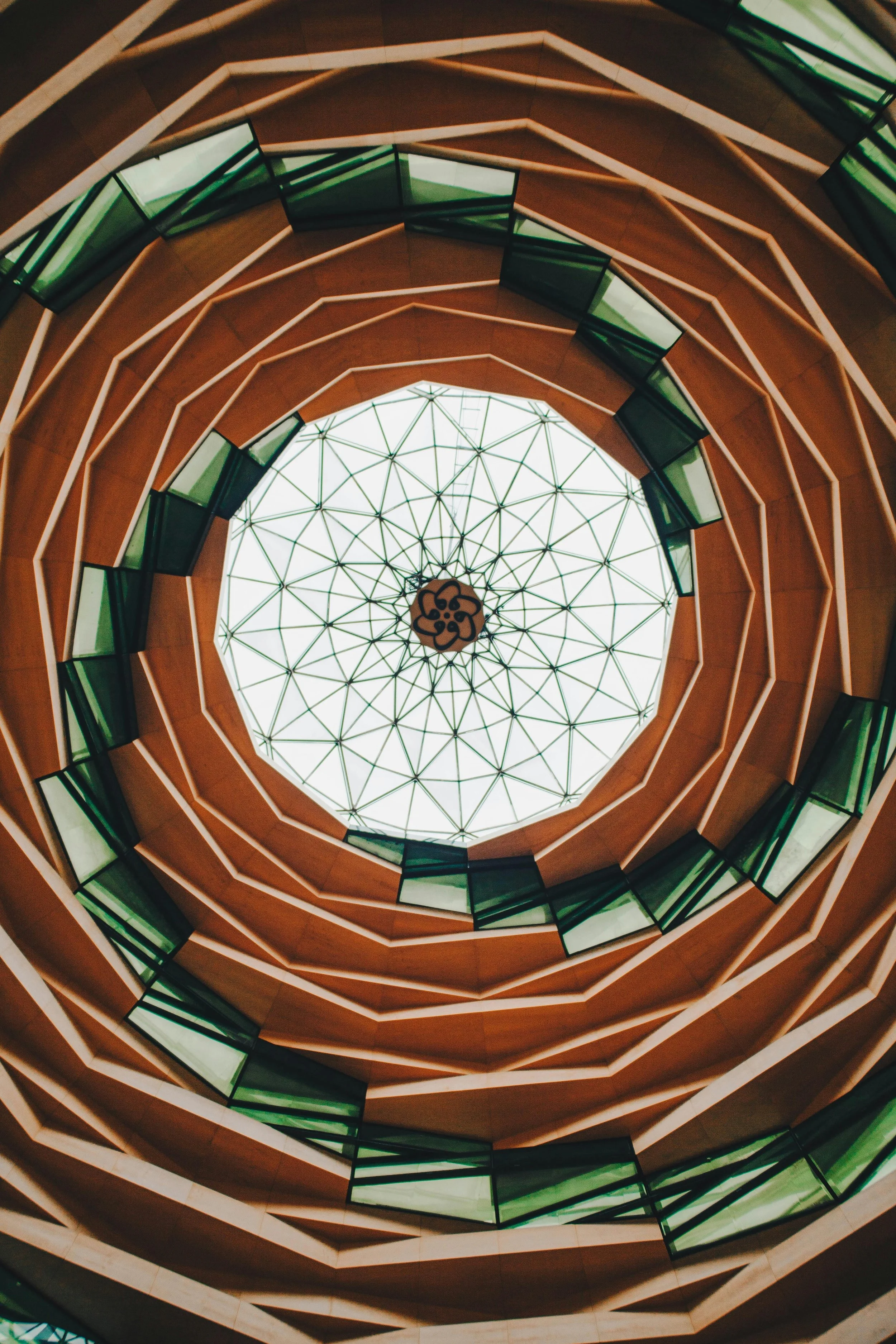 View of a spiraling multi-story staircase seen from below, with green-tinted glass railings and an intricate glass ceiling at the top resembling a geometric pattern.