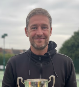 A smiling man with gray hair and beard holding a trophy outdoors, wearing a black hoodie.