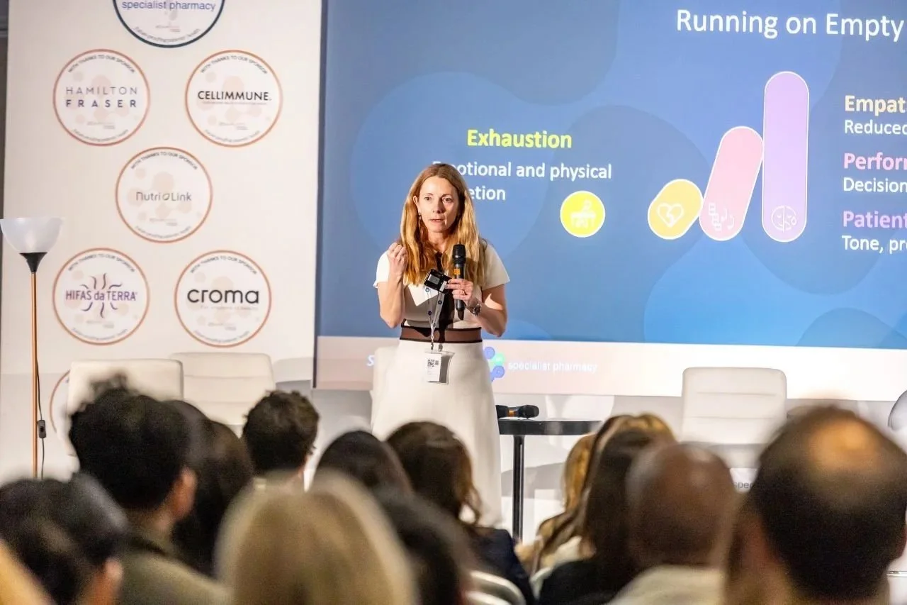 A woman is giving a presentation on a stage at a conference or seminar, holding a microphone. Behind her, a large screen displays a slide with the title 'Running on Empty' and points about exhaustion and emotional and physical fatigue. The audience is seated and listening, and there is a panel of sponsor logos on the left side of the stage.