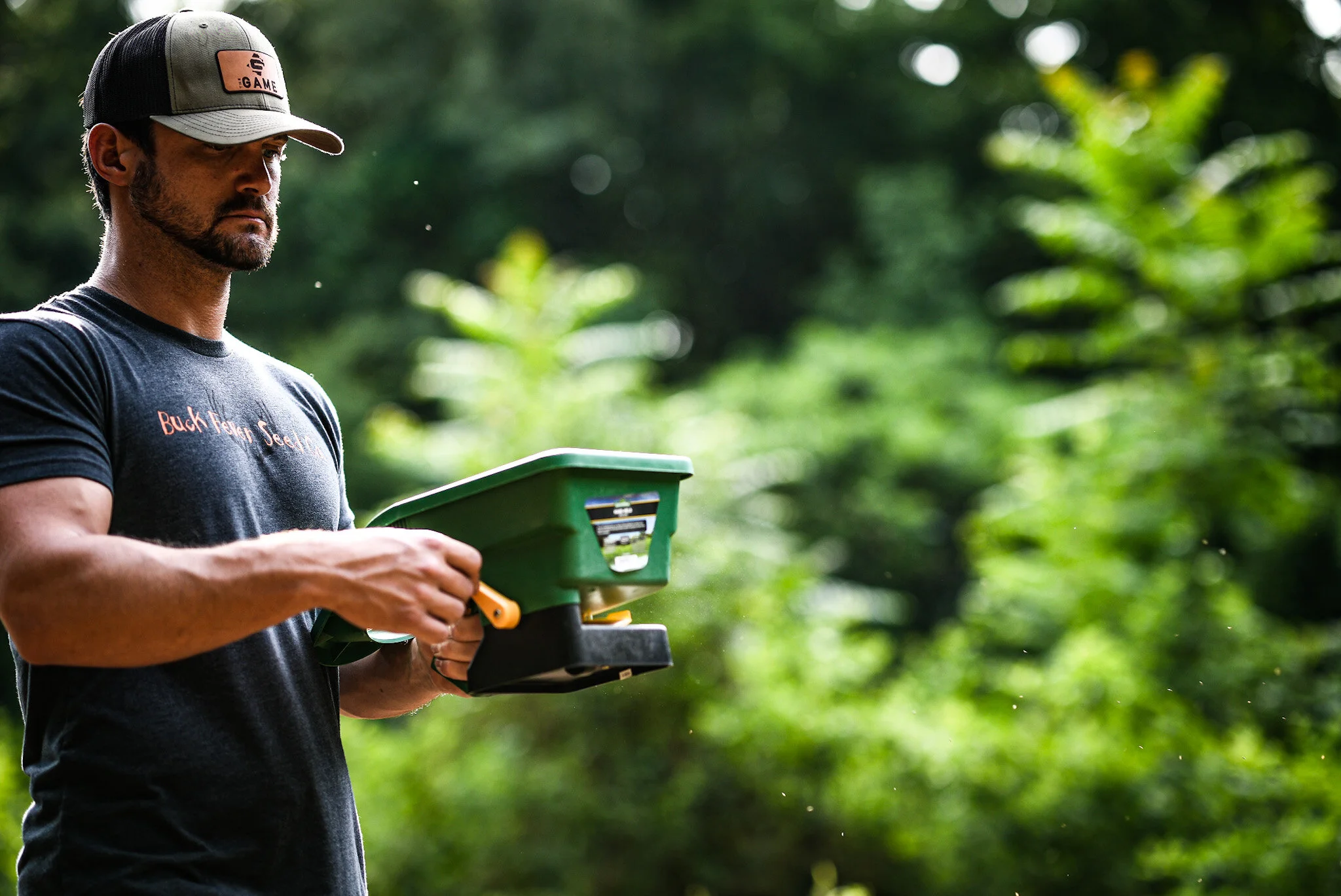 A man with a beard wearing a gray baseball cap and a black T-shirt reading 'Buck Fedge' stands outdoors in a forest setting, holding a green pest control device.