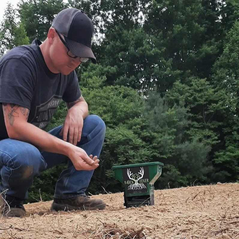 A man wearing glasses, a black cap, and a black T-shirt crouches down near a squirrel feeder shaped like a green bin with deer antlers and a logo reading 'Sick Fever.' The man is outside on a sandy patch with green trees in the background.