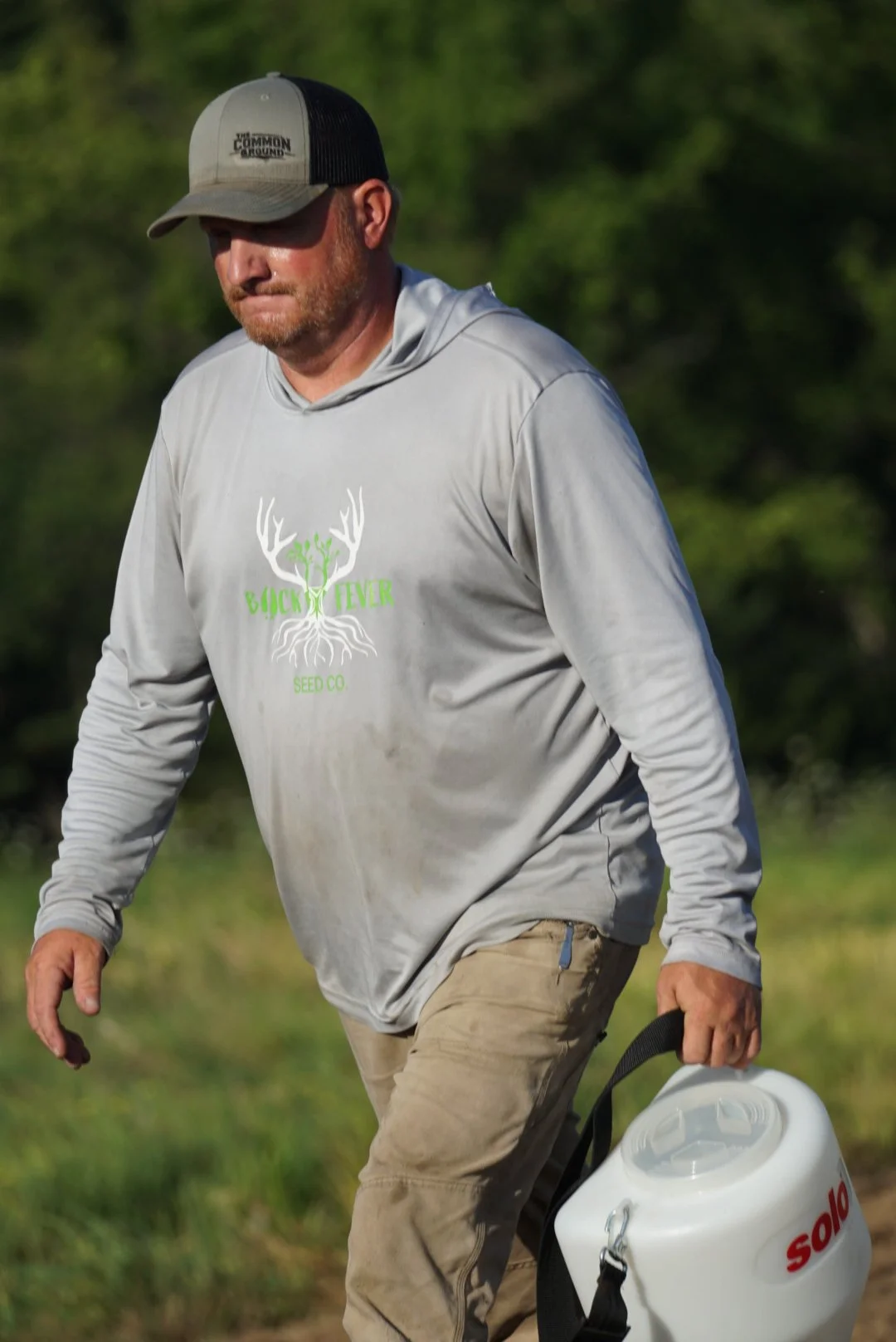 A man walking outdoors wearing a gray hoodie with a green and white logo that says 'Buck Fever Seed Co.' on the front, a black and gray trucker hat, and tan pants. He is holding a white plastic container with a black strap.