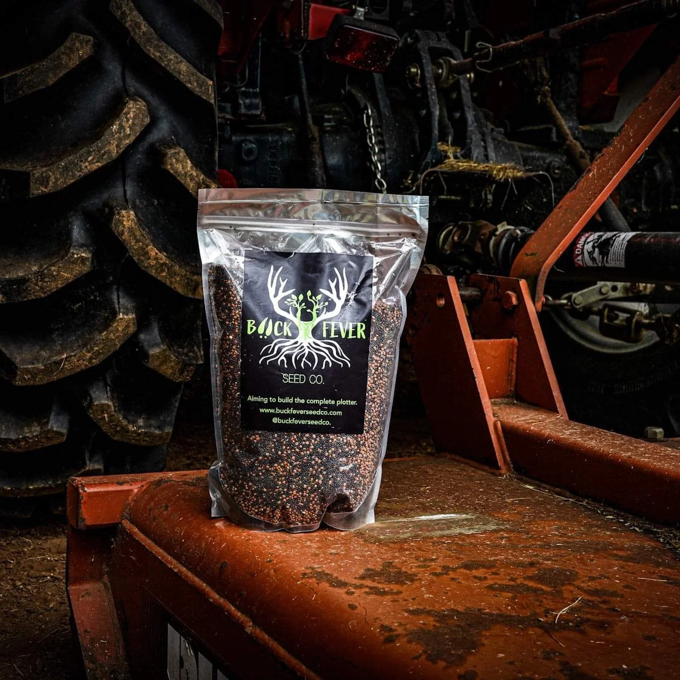 A bag of Buck Fever seed co. planting seed with a graphic of a deer with antlers and roots, placed on a rusty orange metal surface inside a workshop or garage with machinery and large tires in the background.
