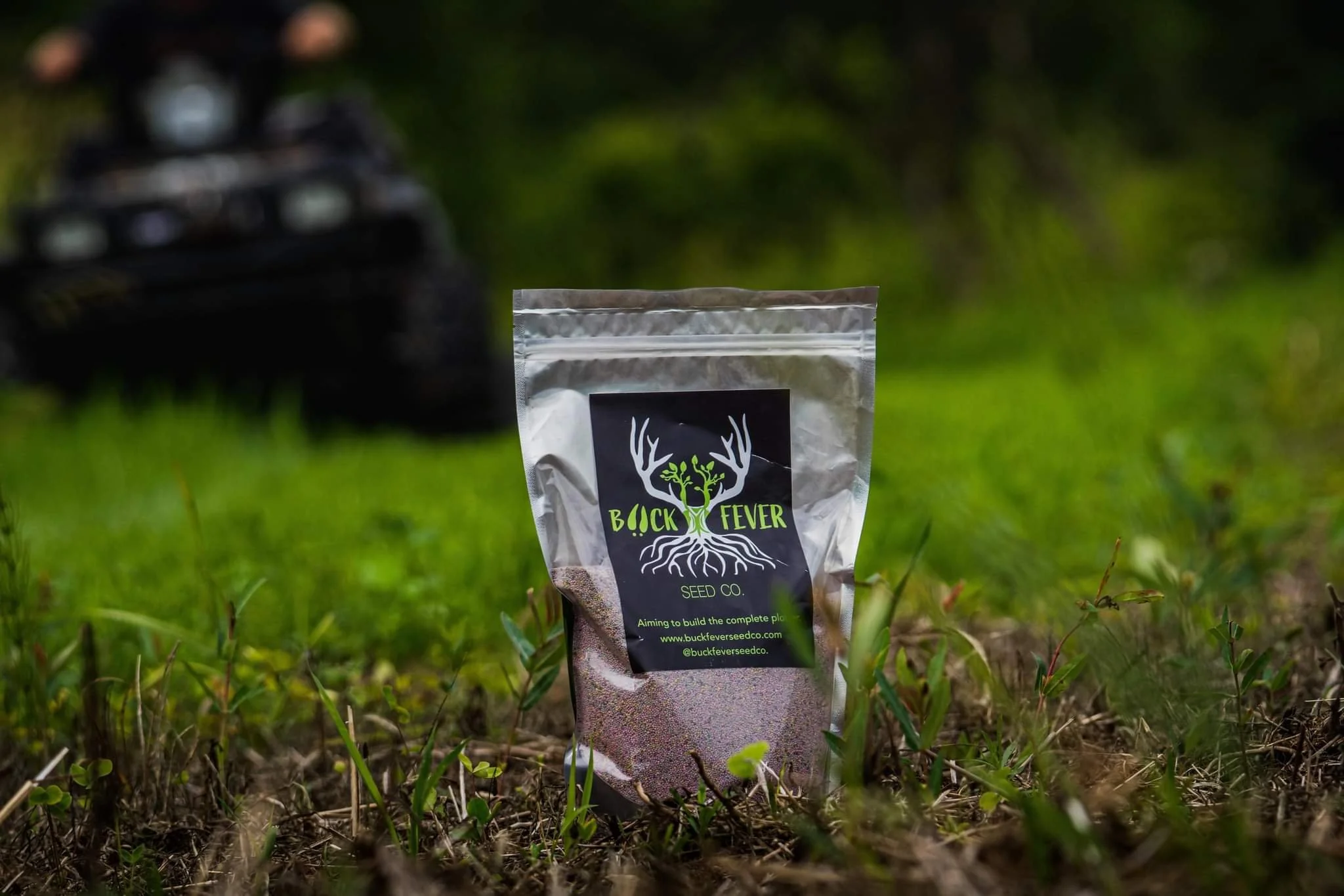 A packet of Buck Fever seed co. seed placed on the grass outdoors with a blurred trail camera in the background.