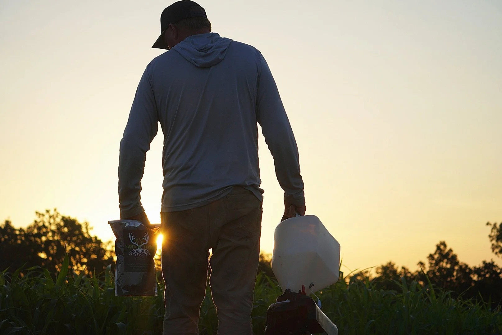 A person standing in a field at sunset, holding a bag of deer feed in one hand and a white gasoline container in the other, with trees in the background.