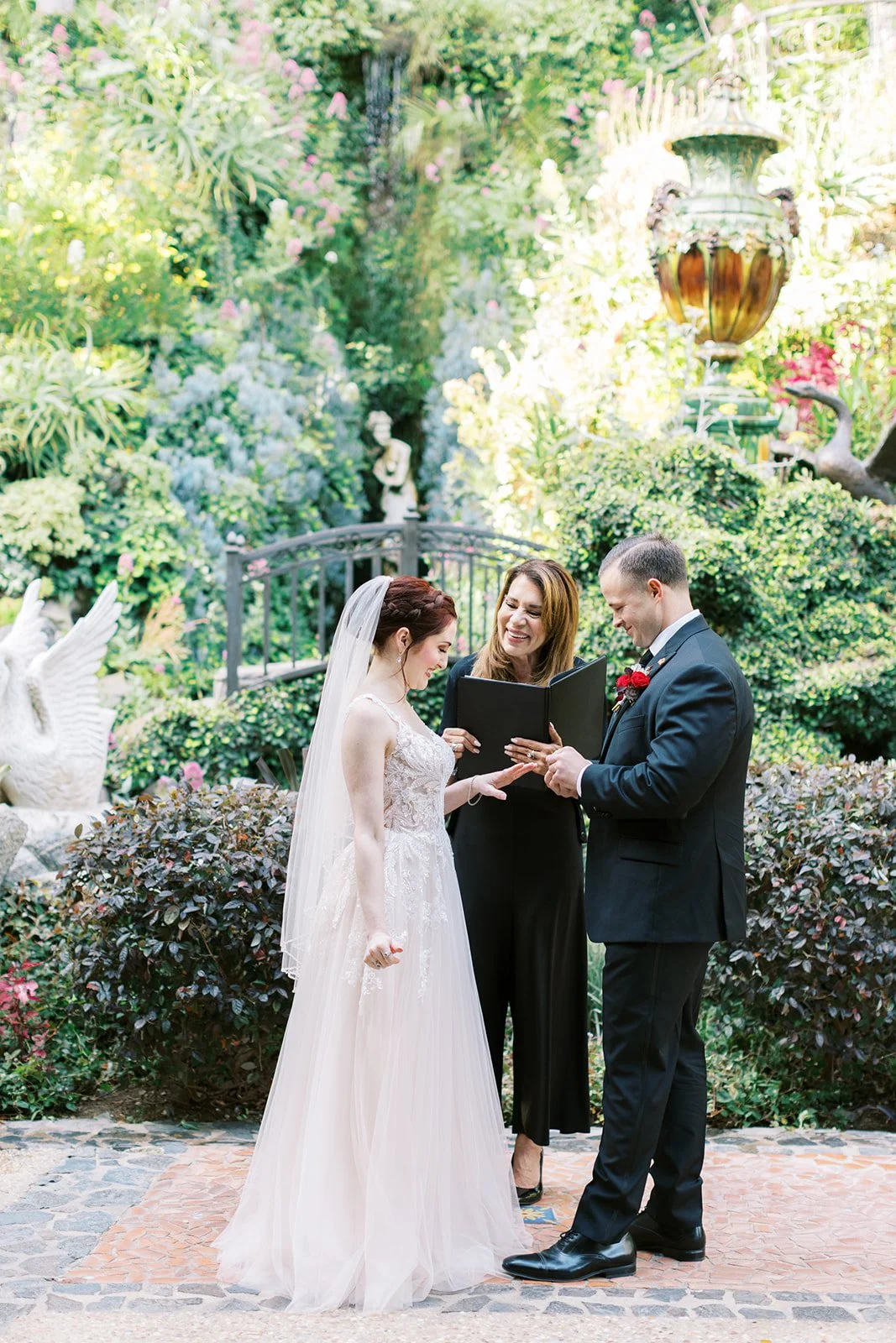 Groom placing the ring on the bride’s finger among the statues and foliage of the Houdini Estate gardens