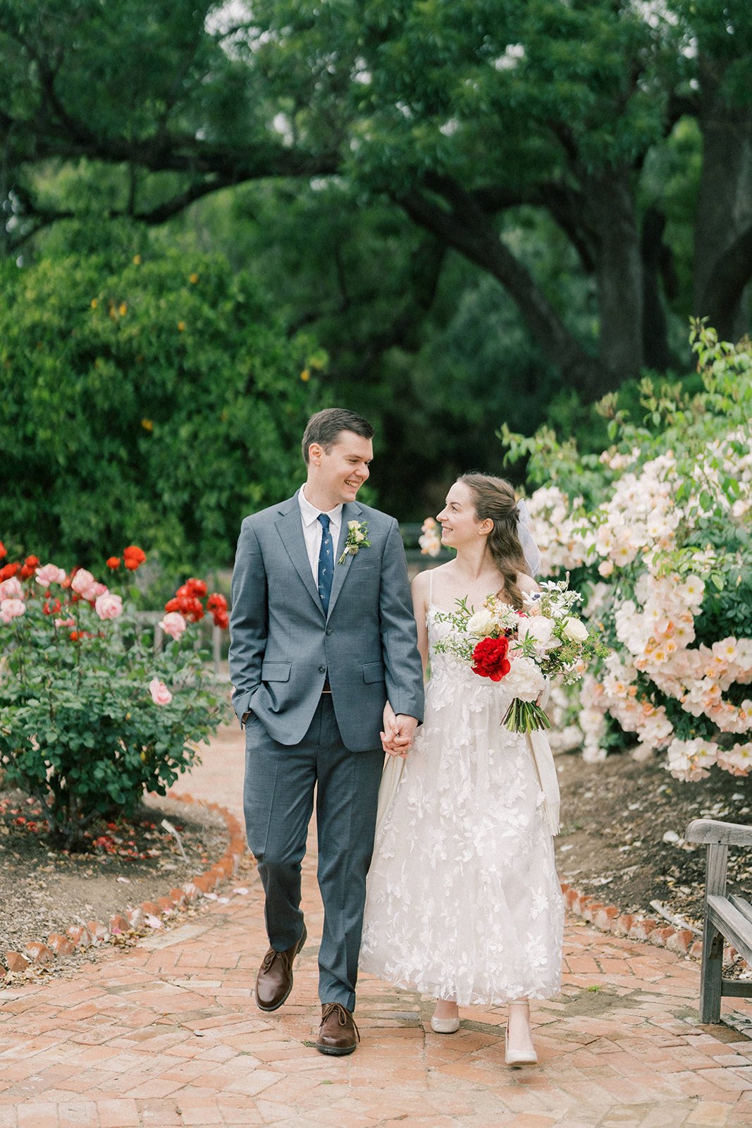 Bride and groom walking hand in hand through the Fullerton Arboretum, gazing at each other as she holds her bouquet