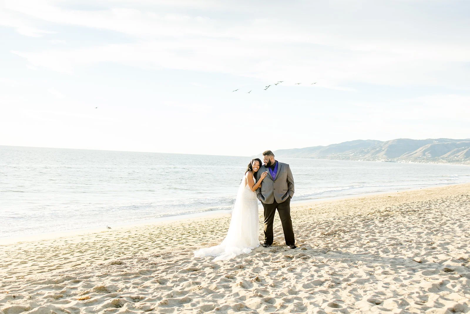 Bride leaning on the groom’s shoulder, both laughing, on the sand at Point Dume in Malibu