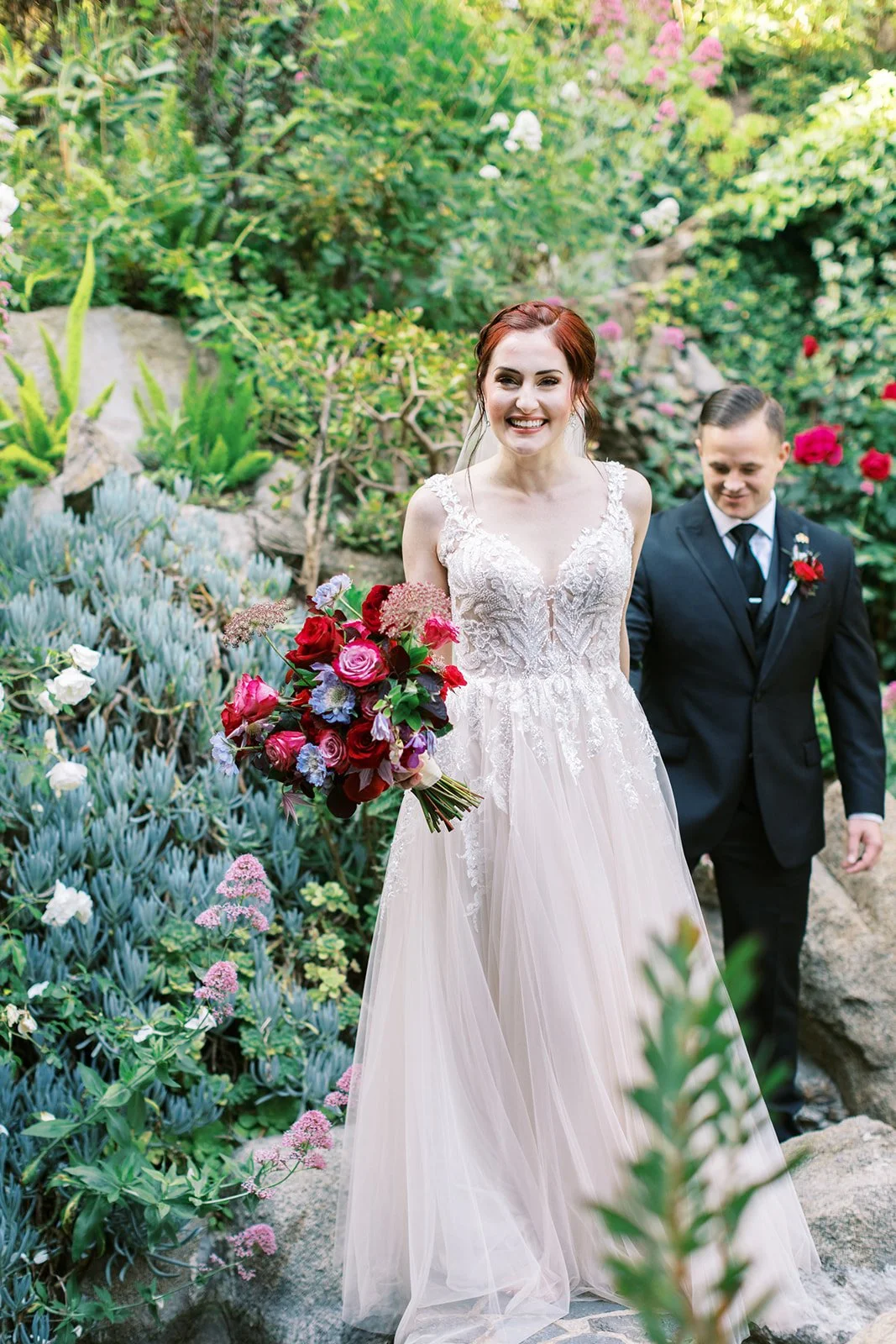 Smiling bride carrying her bouquet along a garden path, the groom walking just behind her