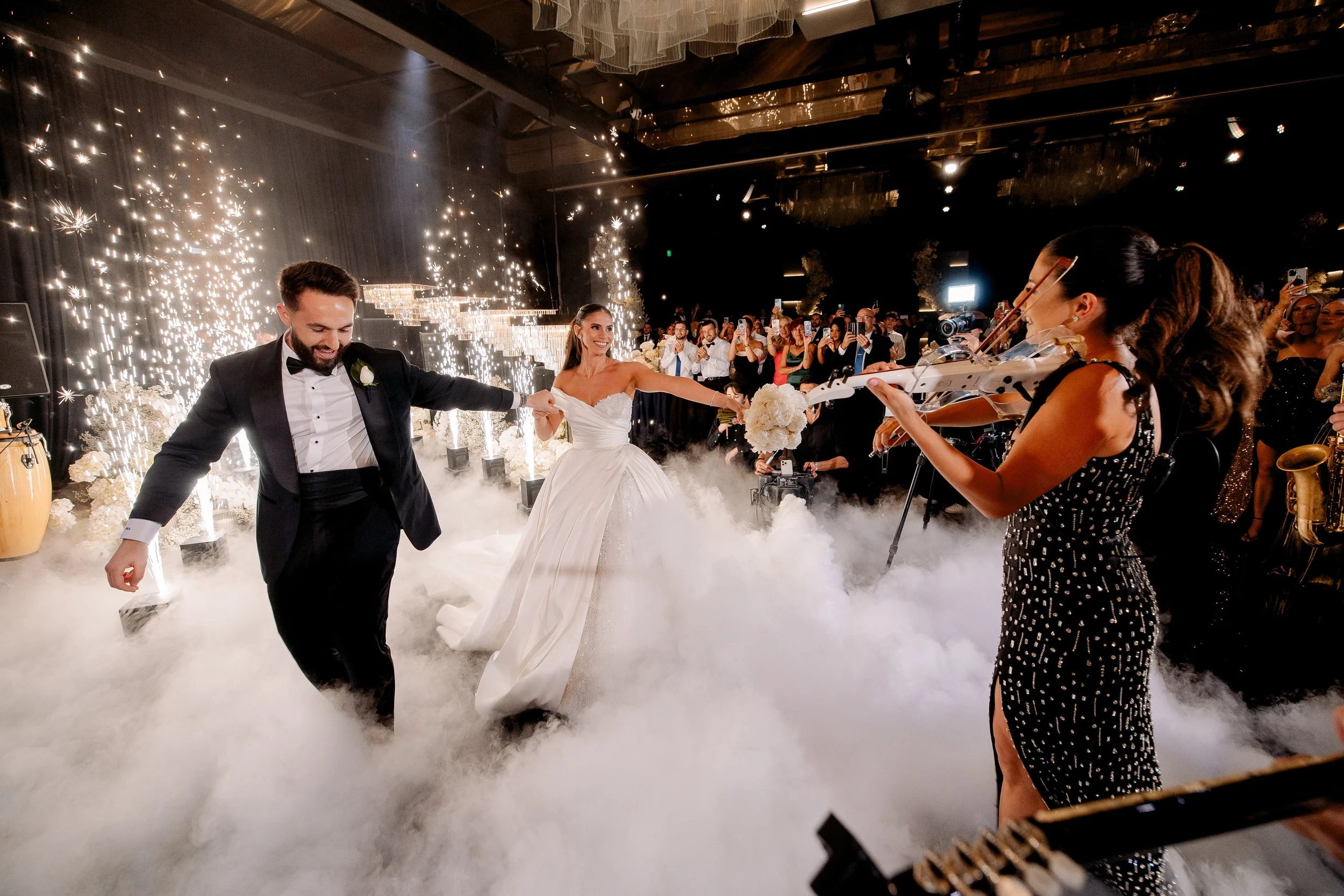 A bride and groom dancing at their wedding reception with sparklers and smoke effects, while a woman plays the violin nearby, and guests watch and take photos.