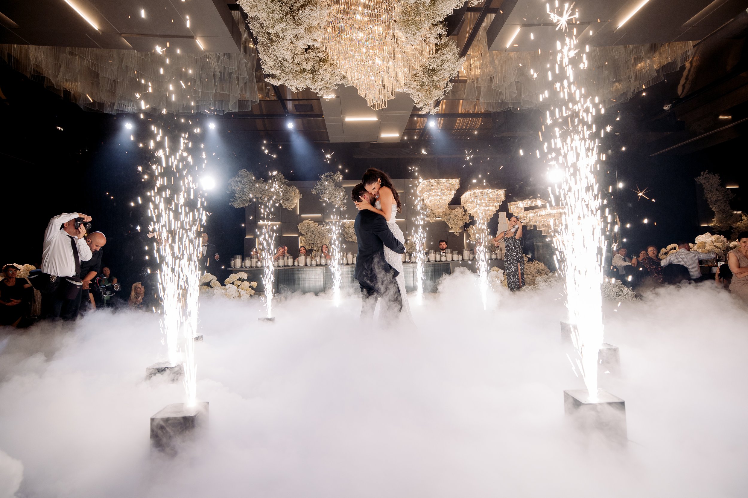 A bride and groom share a dance surrounded by confetti cannons shooting fireworks, with fog covering the floor at their wedding reception.
