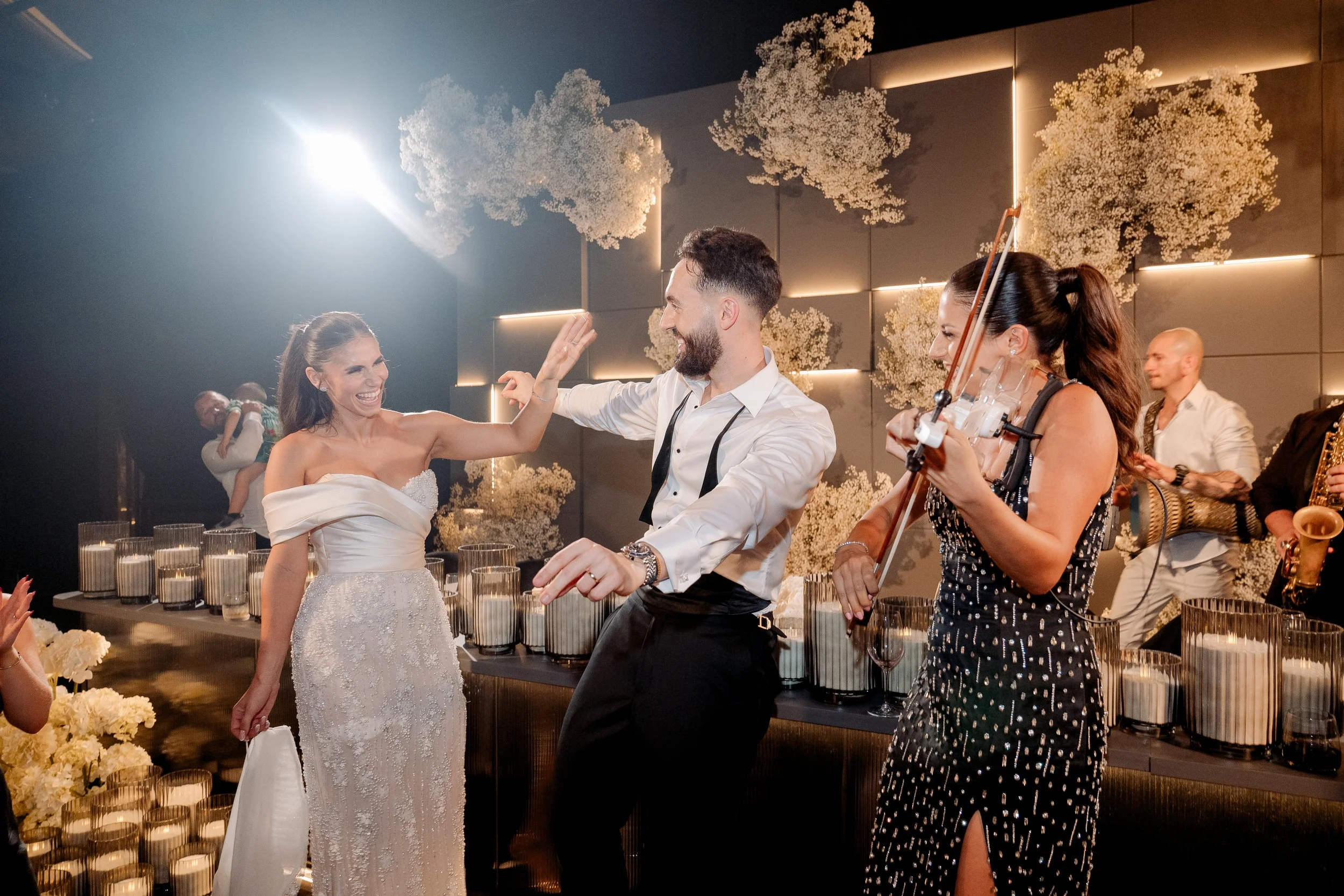 A joyful wedding celebration with a bride in a strapless wedding dress, a groom in a white shirt and black pants dancing, and a violinist in a black dress. The background features a wall with illuminated floral decorations and guests enjoying the mus