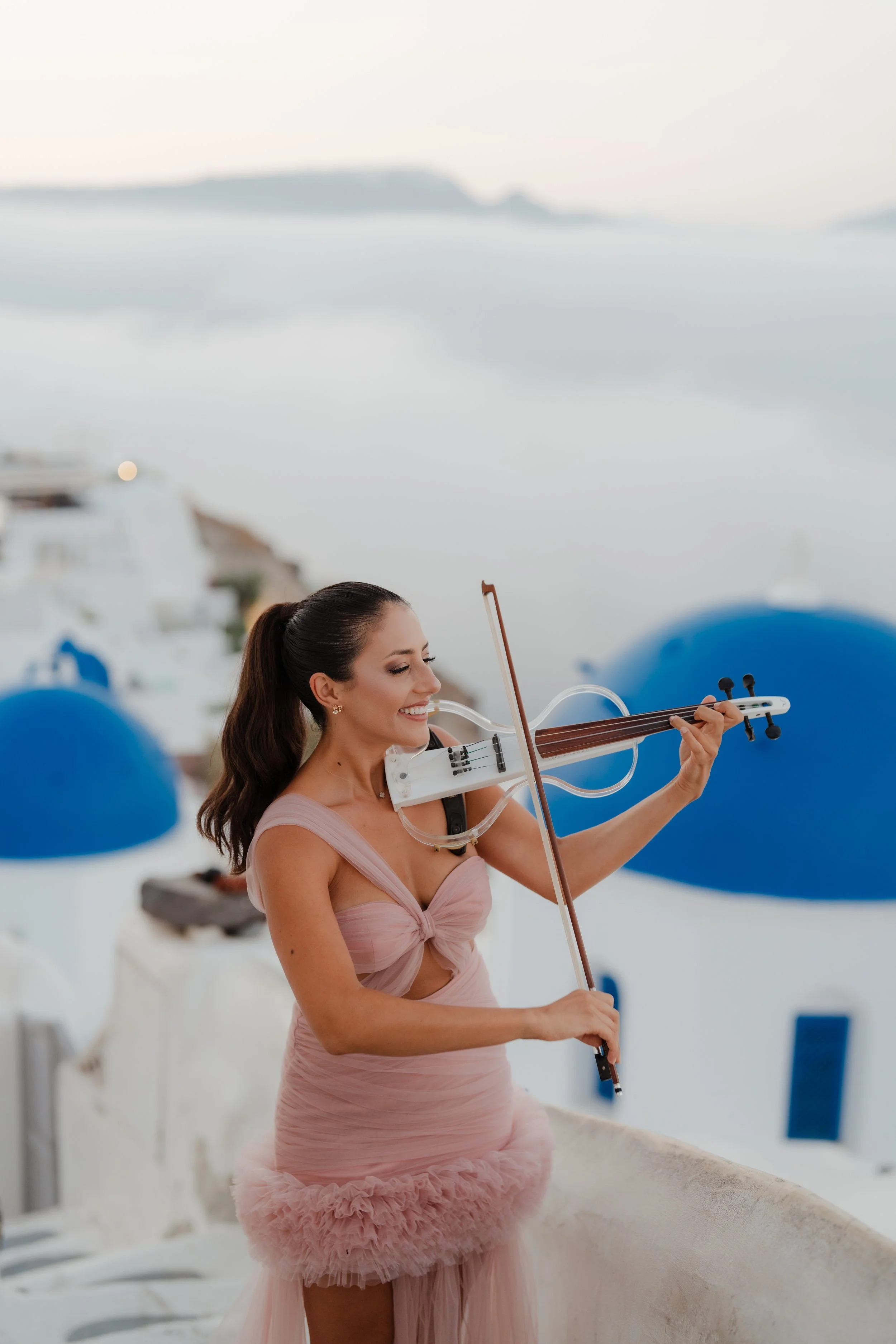 A woman in a pink dress playing a violin on a rooftop with white and blue buildings and a cloudy sky in the background.