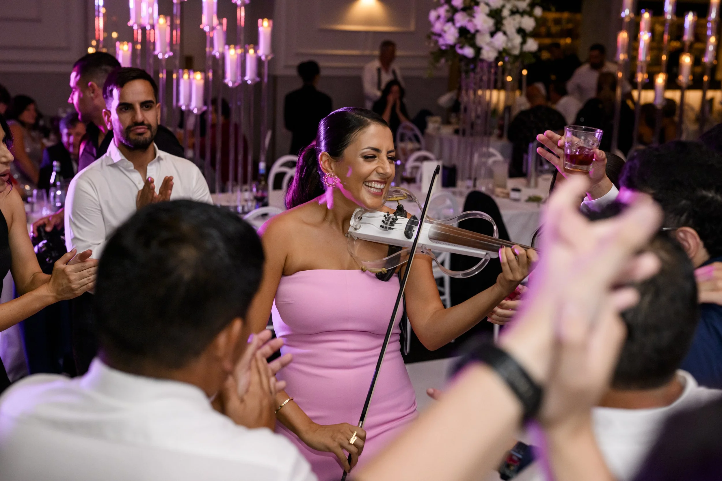 A woman in a pink dress plays a violin and enjoys the music at a lively celebration with friends, with colorful candles and floral arrangements in the background.