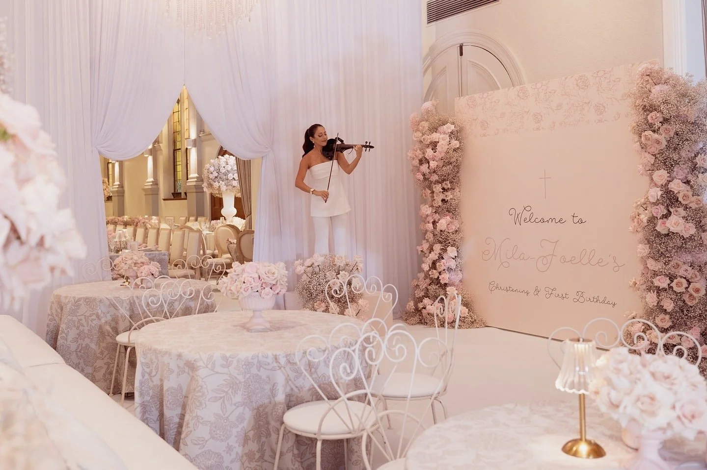 A woman playing the violin in a pastel pink and white decorated event space, with round tables, floral centerpieces, and a large welcome sign for Mila-Faelle's christening and first birthday.