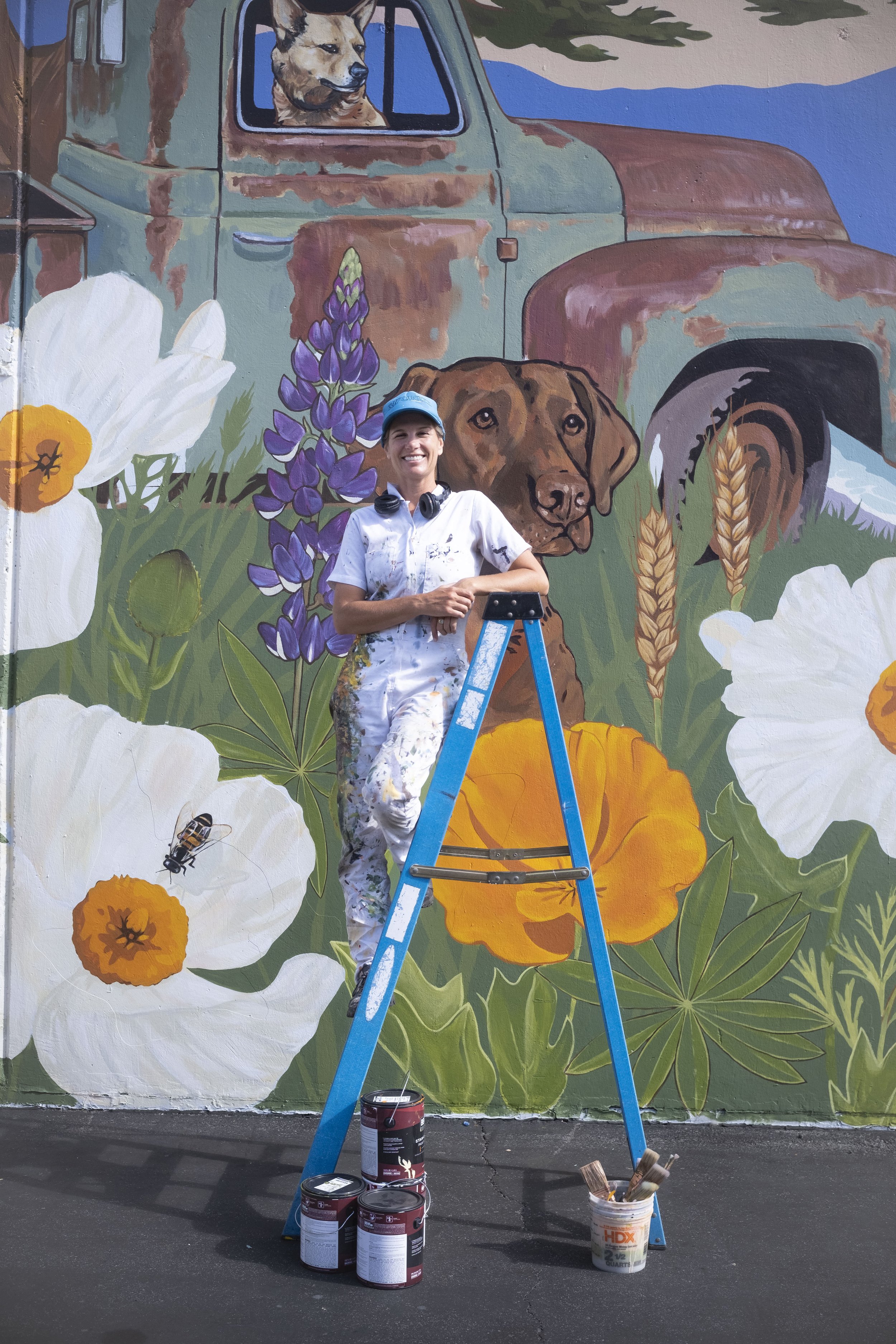 Santa Cruz / Aptos, CA painter, muralist, and artist standing on a ladder, smiling, in front of one of her local murals with flowers, dogs, and an old rusty truck in it