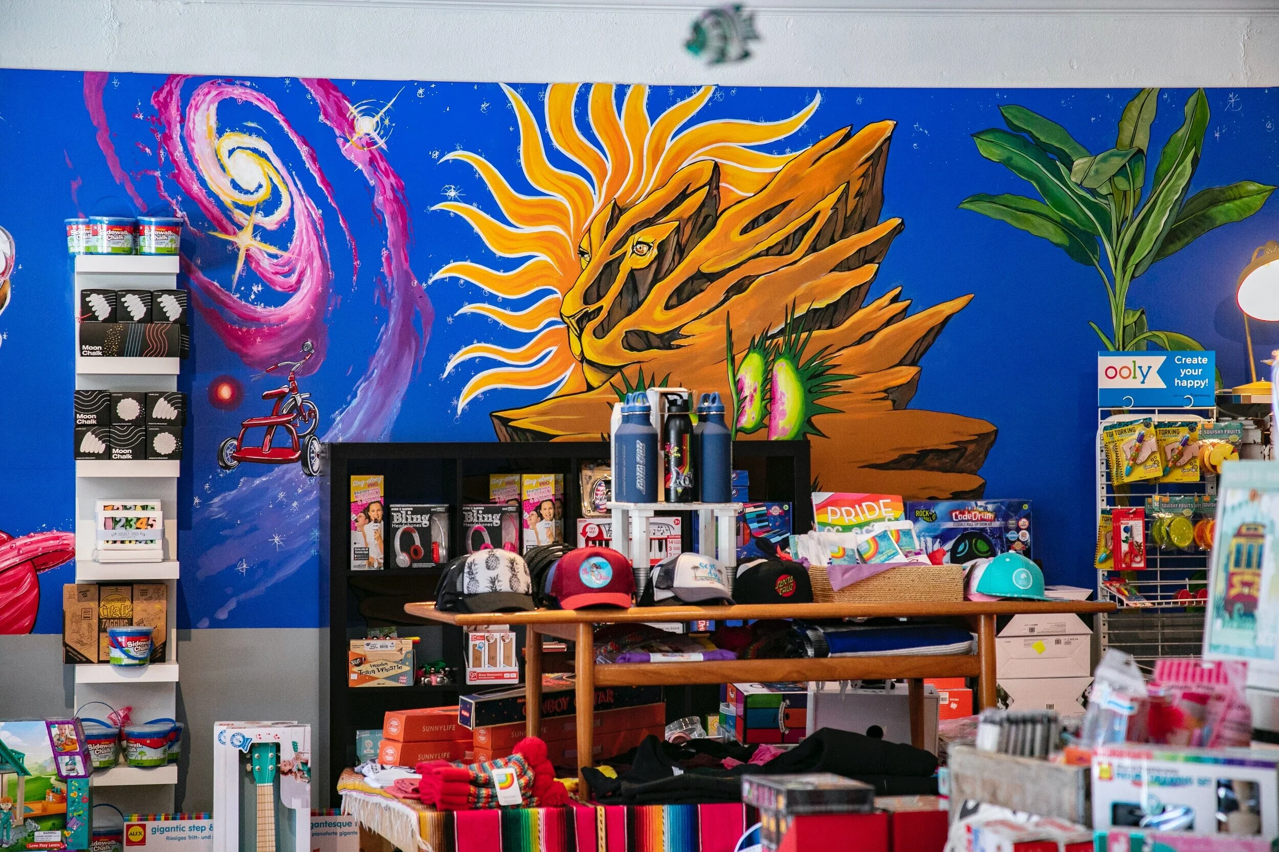 Childish Toy Shop indoor Mural in Santa Cruz, California full of space theme whimsical creatures, a lion faced sun, a vortex, a tricycle, plants, and more by local painter, Brittany Costanzo