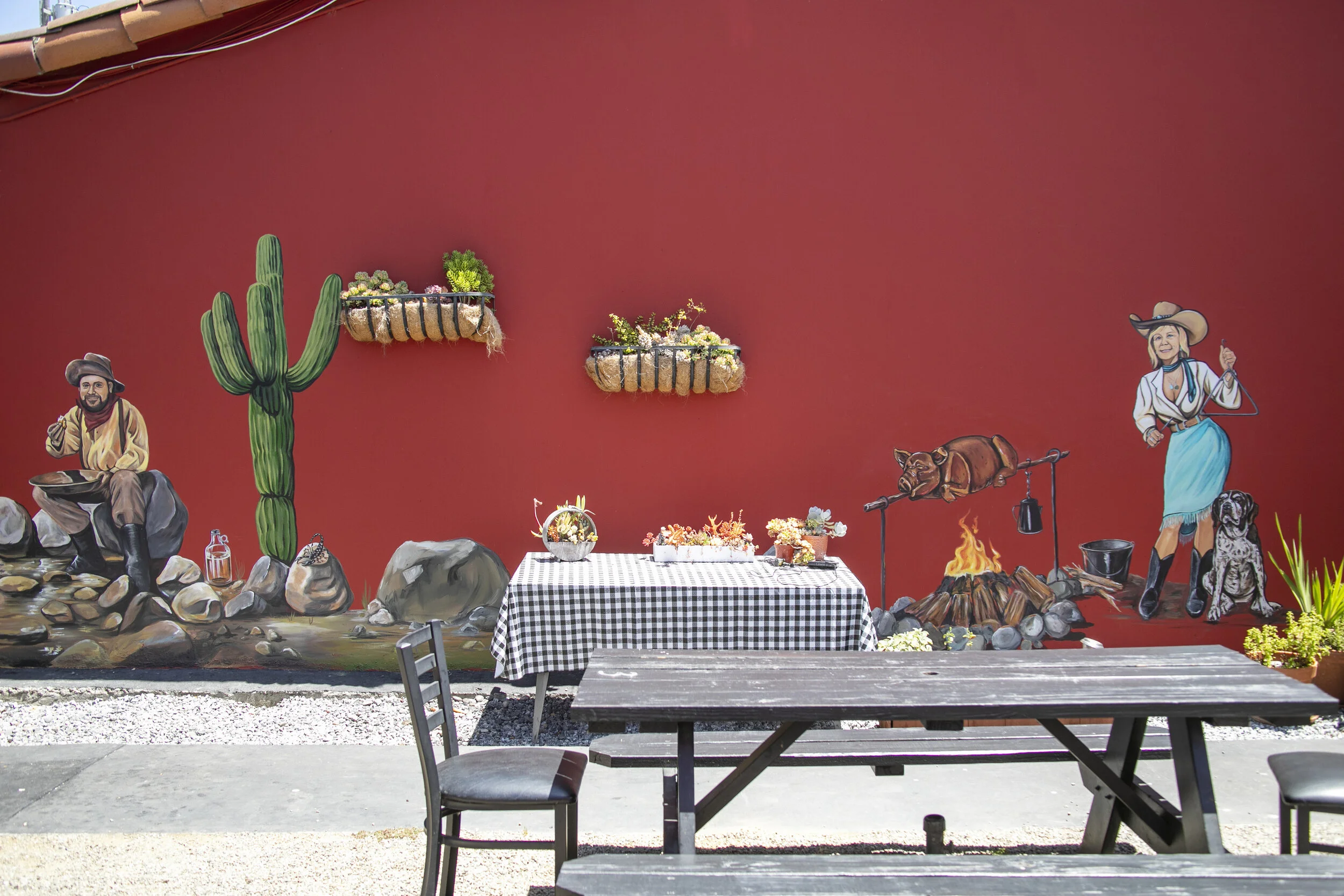 Holy Smokes BBQ & Catering Mural of cowboys and cowgirls in western attire, roasting a pig over an open fire with a do, surrounded by cactus, rocks, and a western scene on a red wall in Santa Cruz, California by Brittany Costanzo of Costanzo Creative