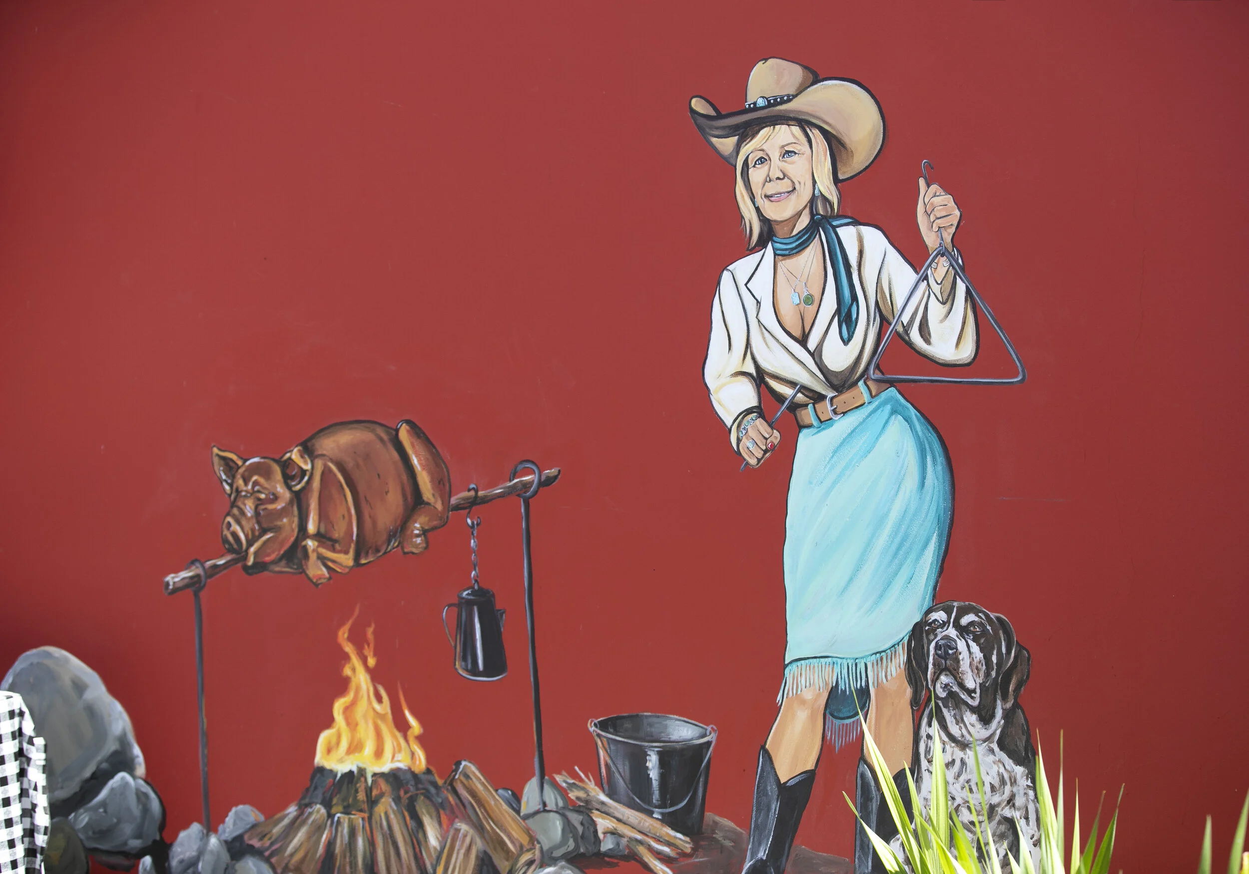 Holy Smokes BBQ & Catering Mural of a cowgirl in western attire, roasting a pig over an open fire with a dog in Santa Cruz, California by Brittany Costanzo