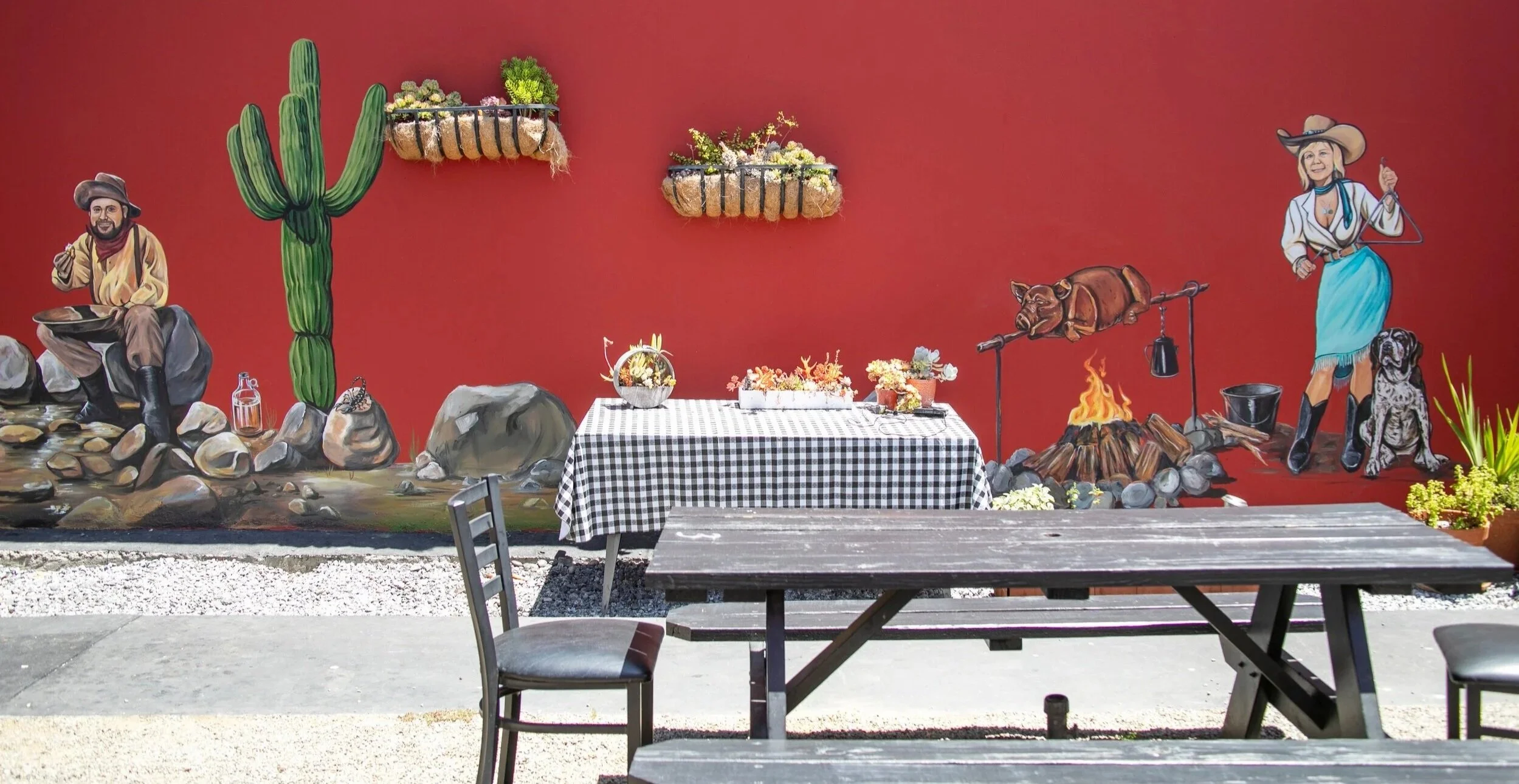 Holy Smokes BBQ & Catering Mural of cowboys and cowgirls in western attire, roasting a pig over an open fire with a dog, cactus, rocks, and other desert wildlife nearby in Santa Cruz, California by Brittany Costanzo
