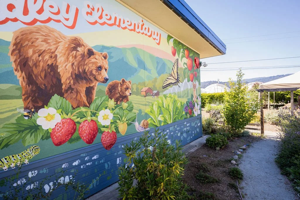 Bradley Elementary School Mural featuring grizzly bears, monarch butterflies, honey bees, strawberries, green hills, red apples, blackberries, and other local flowers, creatures, and plants by Brittany Costanzo Creative 2026