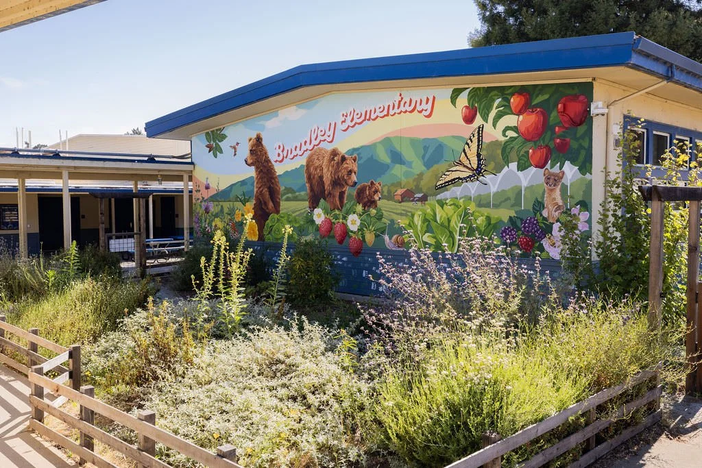 Bradley Elementary School Mural featuring grizzly bears, monarch butterflies, honey bees, strawberries, green hills, red apples, blackberries, and other local flowers, creatures, and plants by Brittany Costanzo Creative 2026