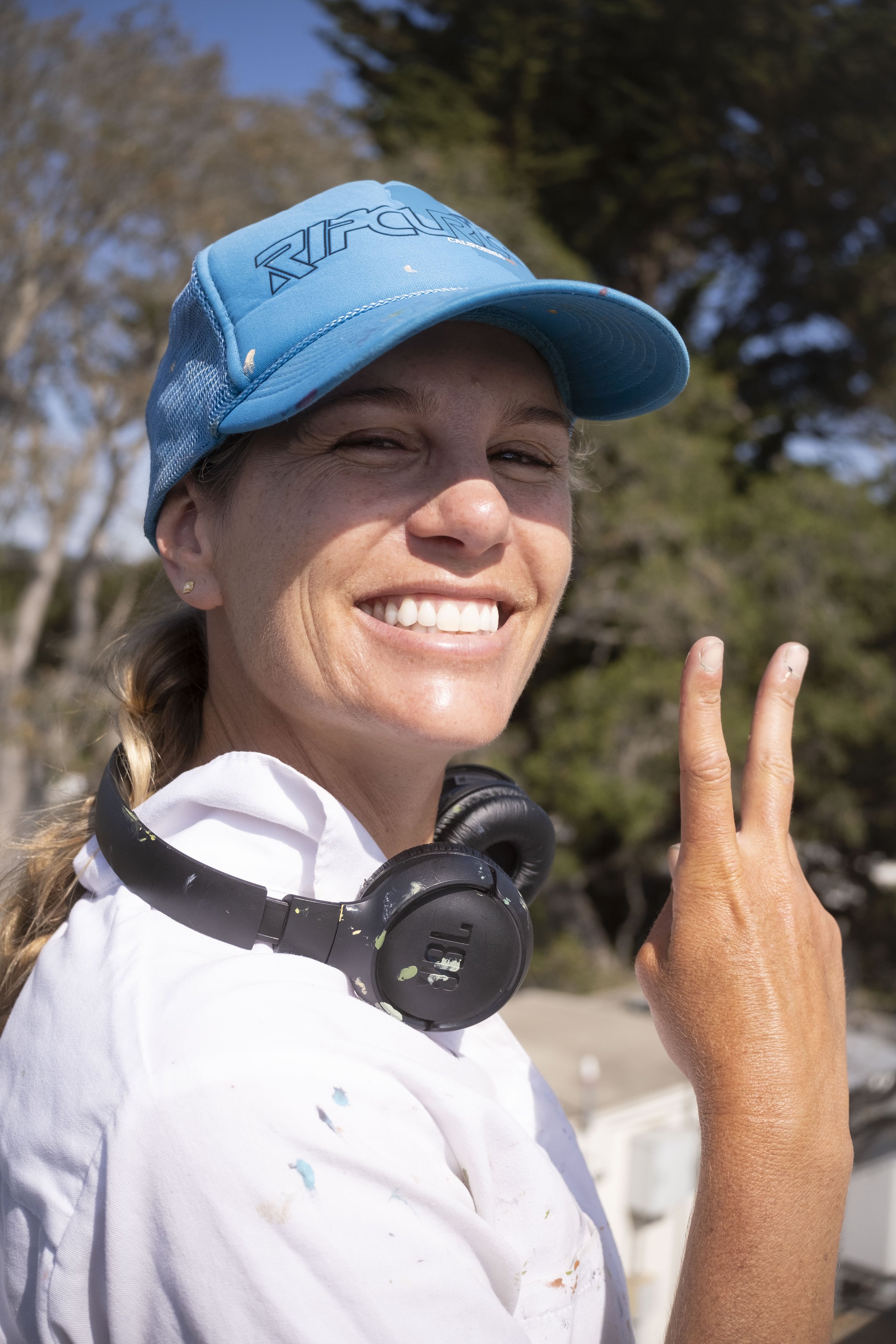 Santa Cruz artist, muralist, painter, Brittany Costanzo, smiling and giving a peace sign with her hand and a Rip Curl trucker hat on