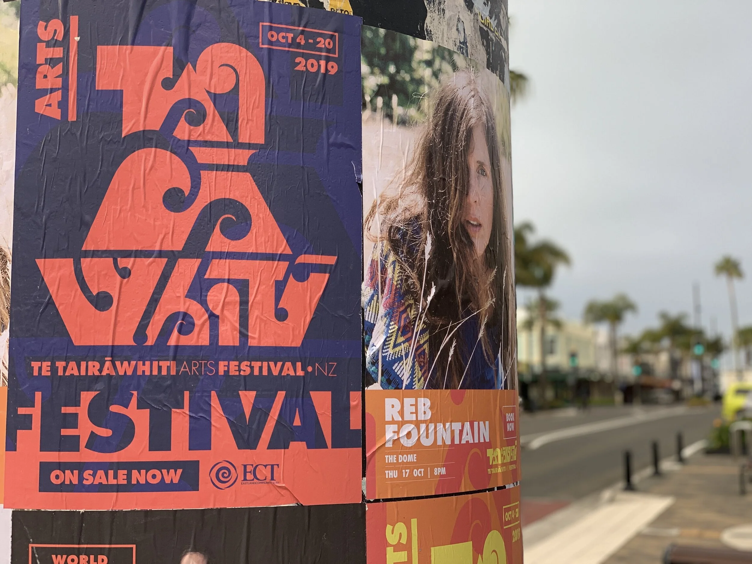 Promotional posters attached to a pole for the Te Tairāwhiti Arts Festival in New Zealand, featuring abstract design and a woman's portrait, with a city street and palm trees in the background.