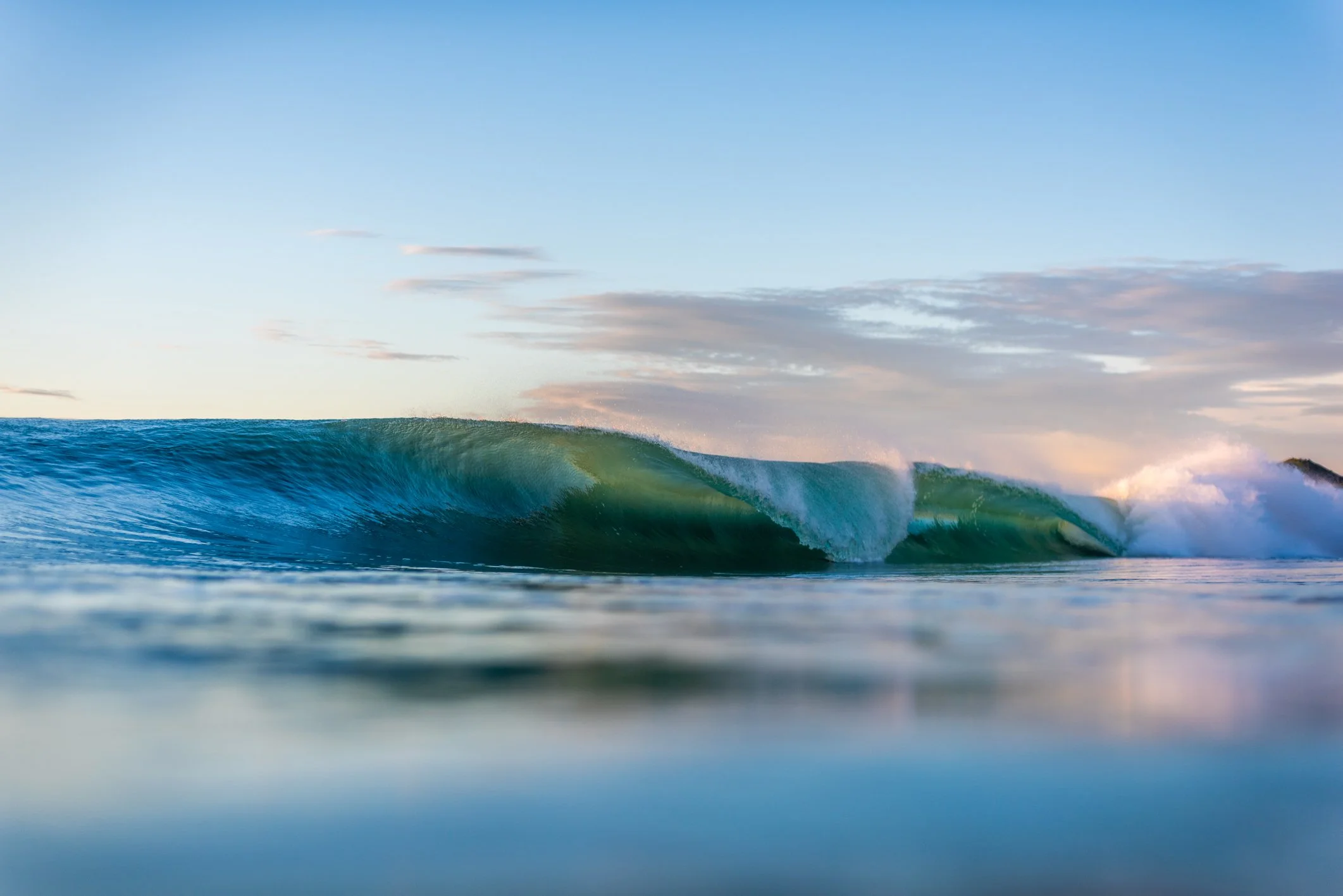 Gentle ocean wave breaking on the shore under a partly cloudy sky at sunset.
