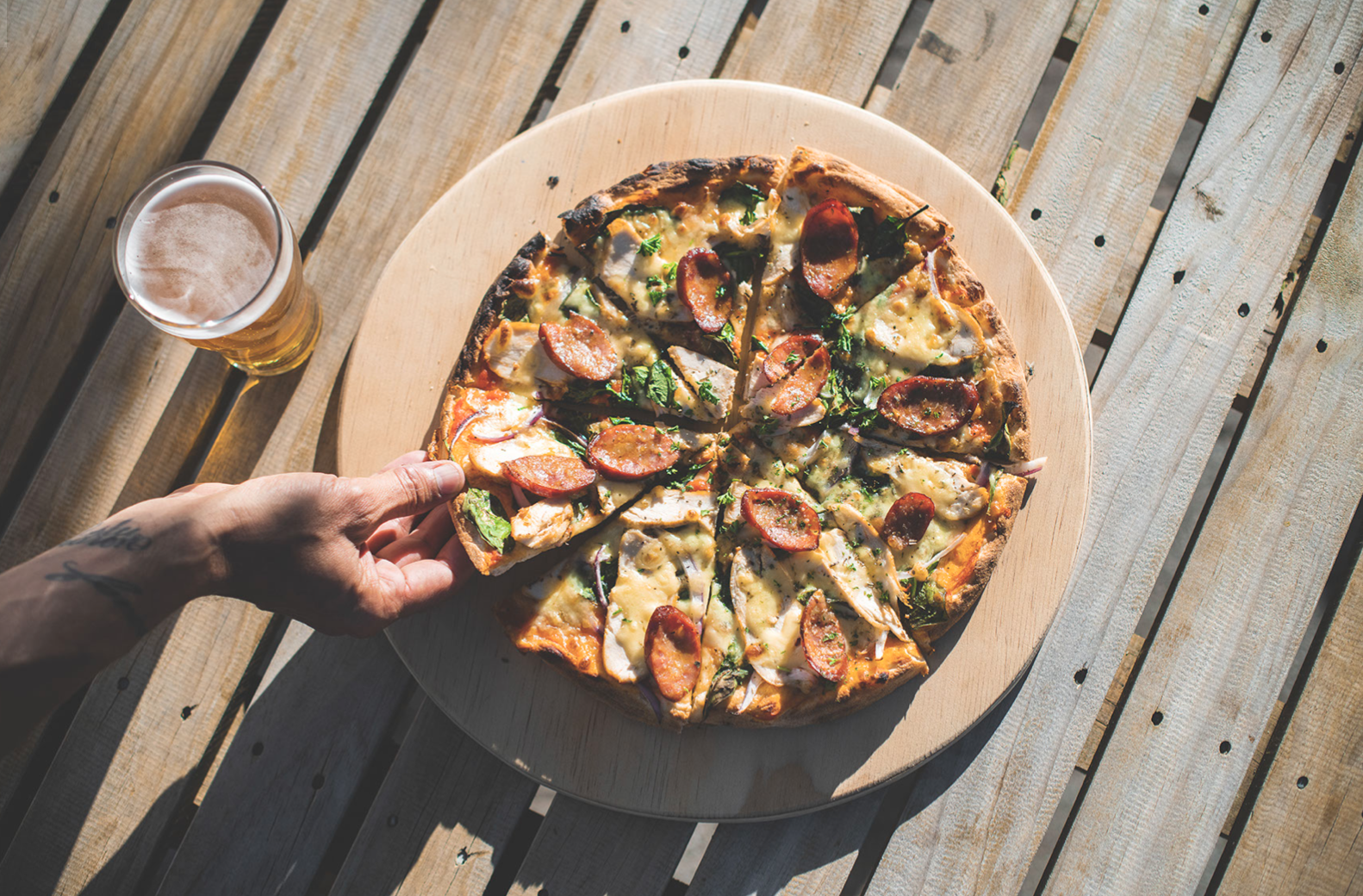 A person with a tattoo on their hand is taking a slice of pizza with pepperoni, vegetables, and cheese from a whole pizza on a round wooden board. There is a glass of beer on the table, which is made of wooden planks.