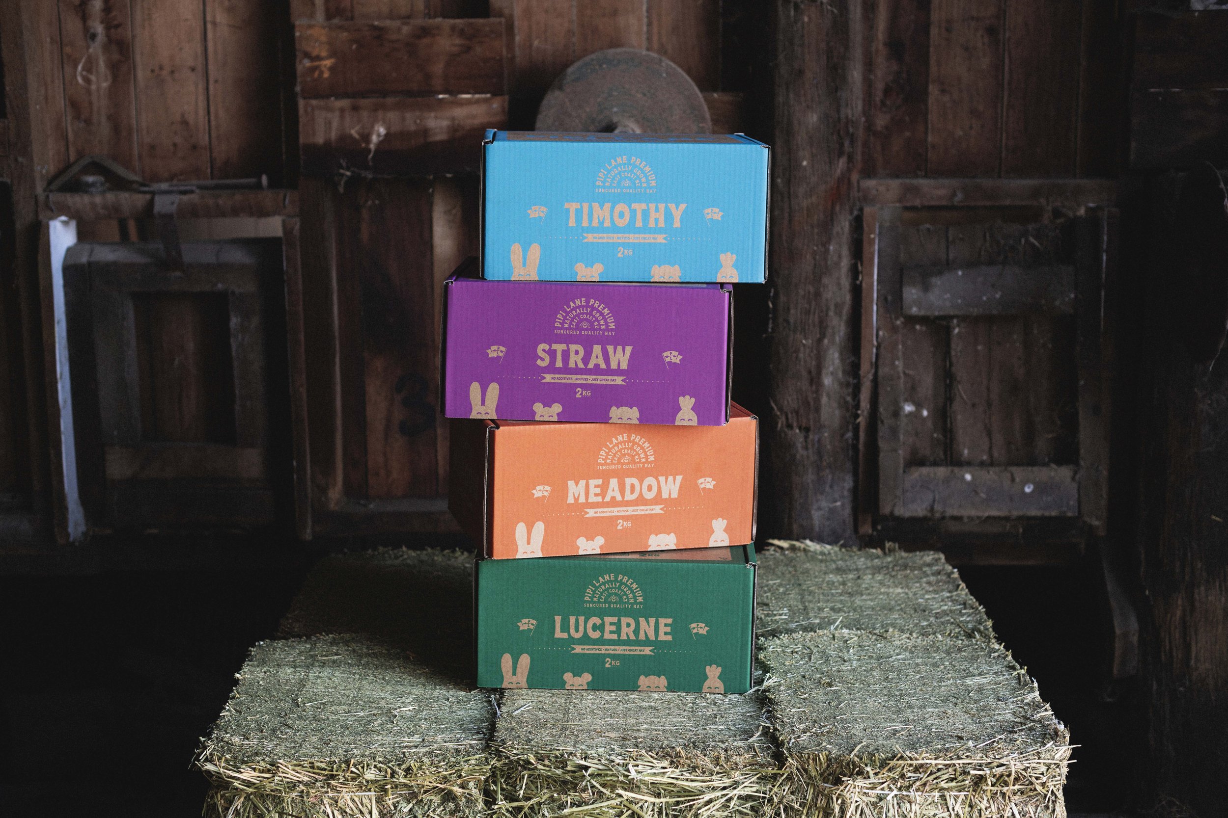 Four colorful boxes stacked on a hay bale inside a rustic wooden barn. The boxes are labeled