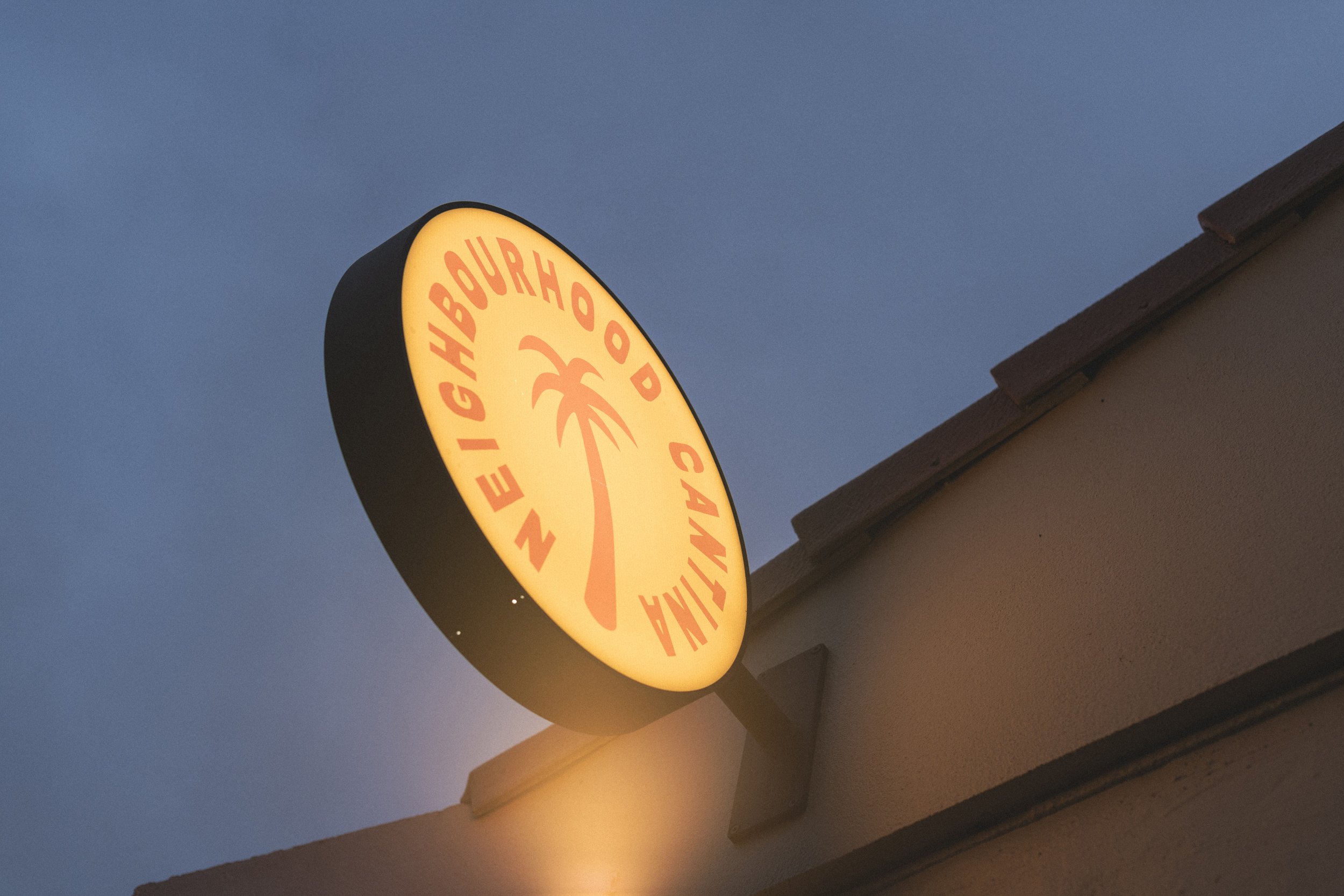 Illuminated sign for Neighbourhood Cantina with a palm tree logo, mounted on a building exterior against a dark sky.