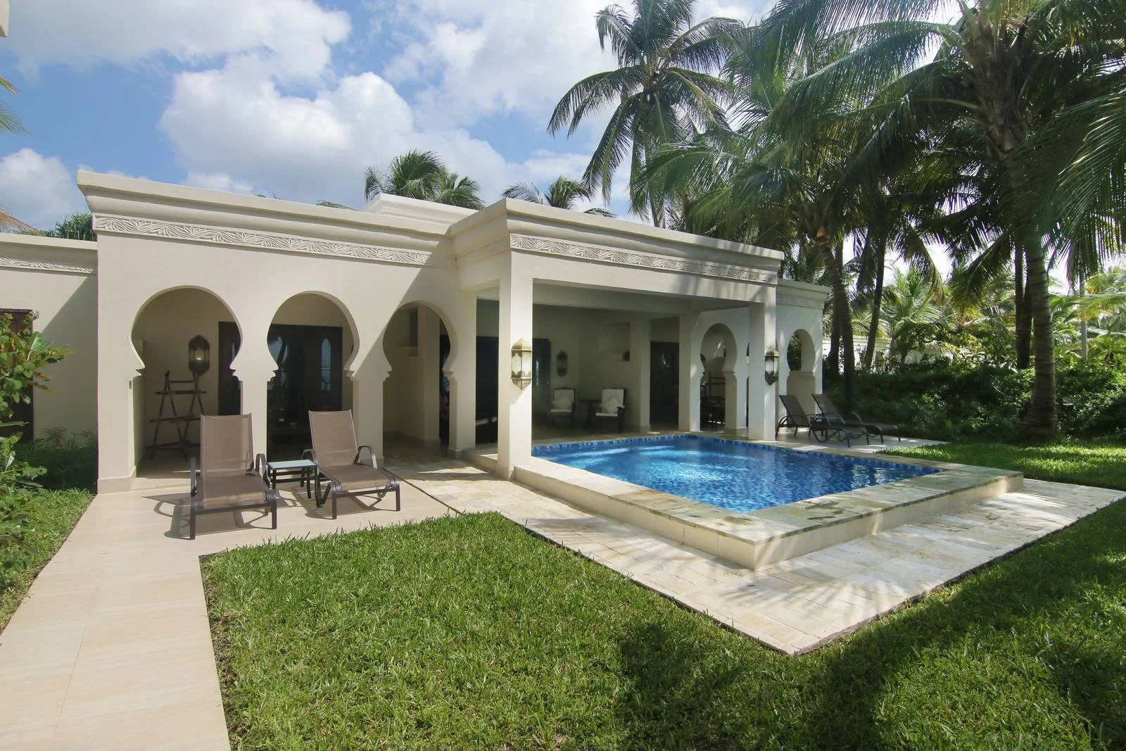 Luxury villa with a private swimming pool surrounded by lush green grass and tall palm trees under a partly cloudy sky.