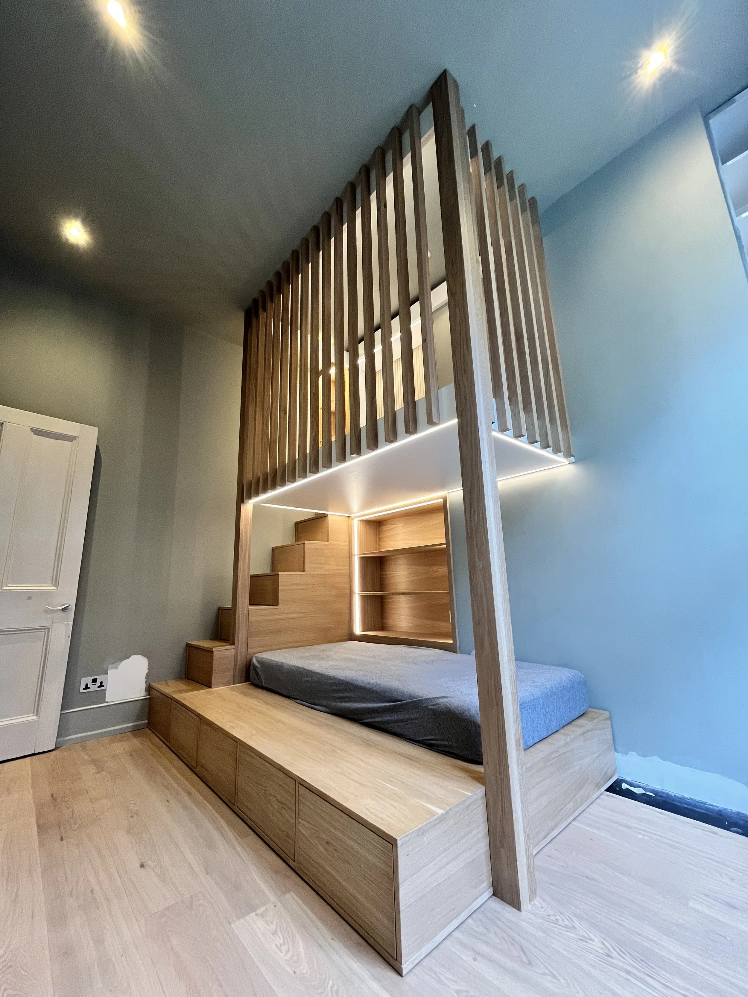 A modern loft bed made of light wood with storage drawers underneath, ceiling lights, and a sleek staircase, in a minimalist bedroom.