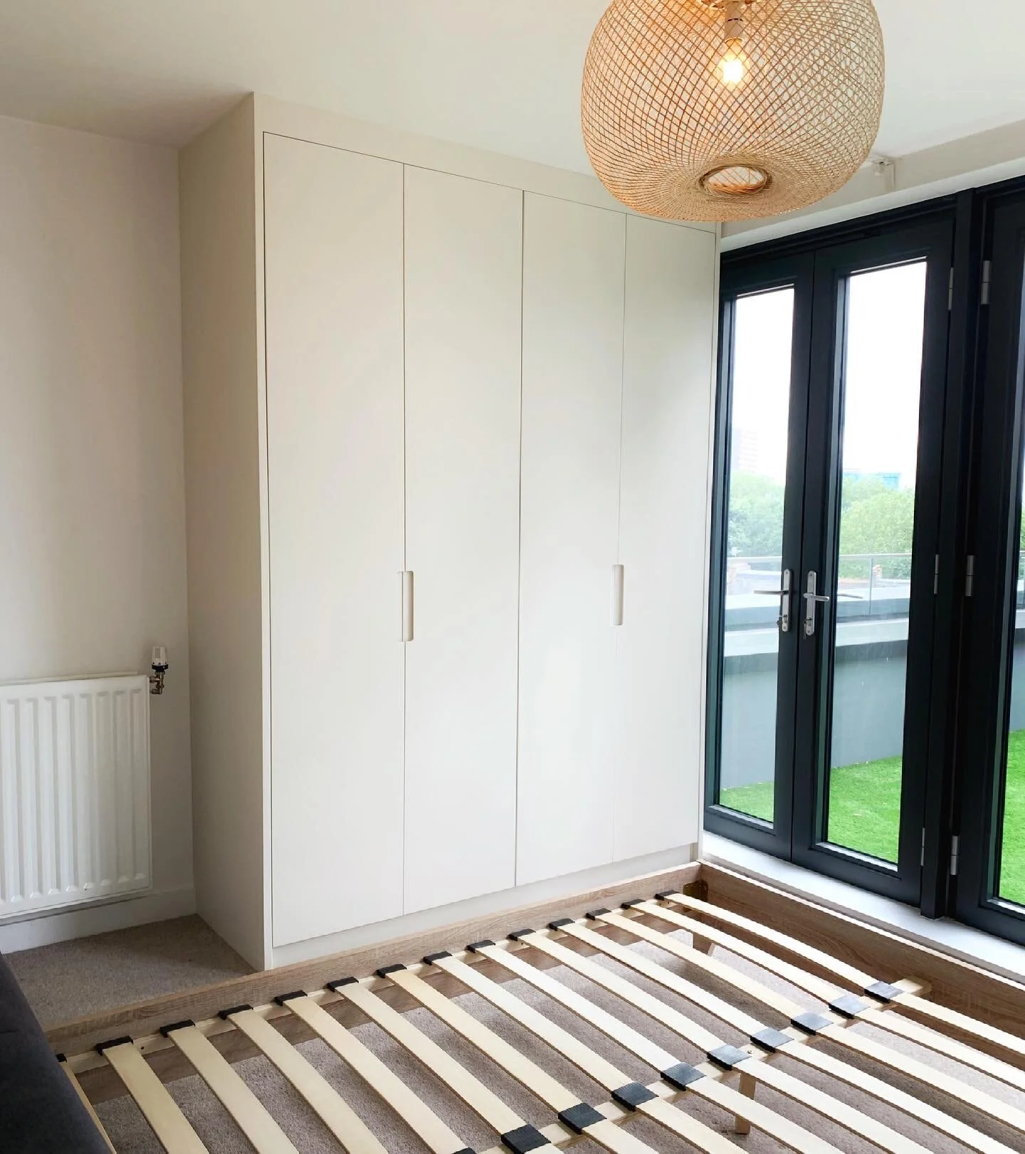 Empty bedroom with a built-in white wardrobe, a hanging woven pendant light, and a bed frame without a mattress next to sliding glass doors leading to a balcony.
