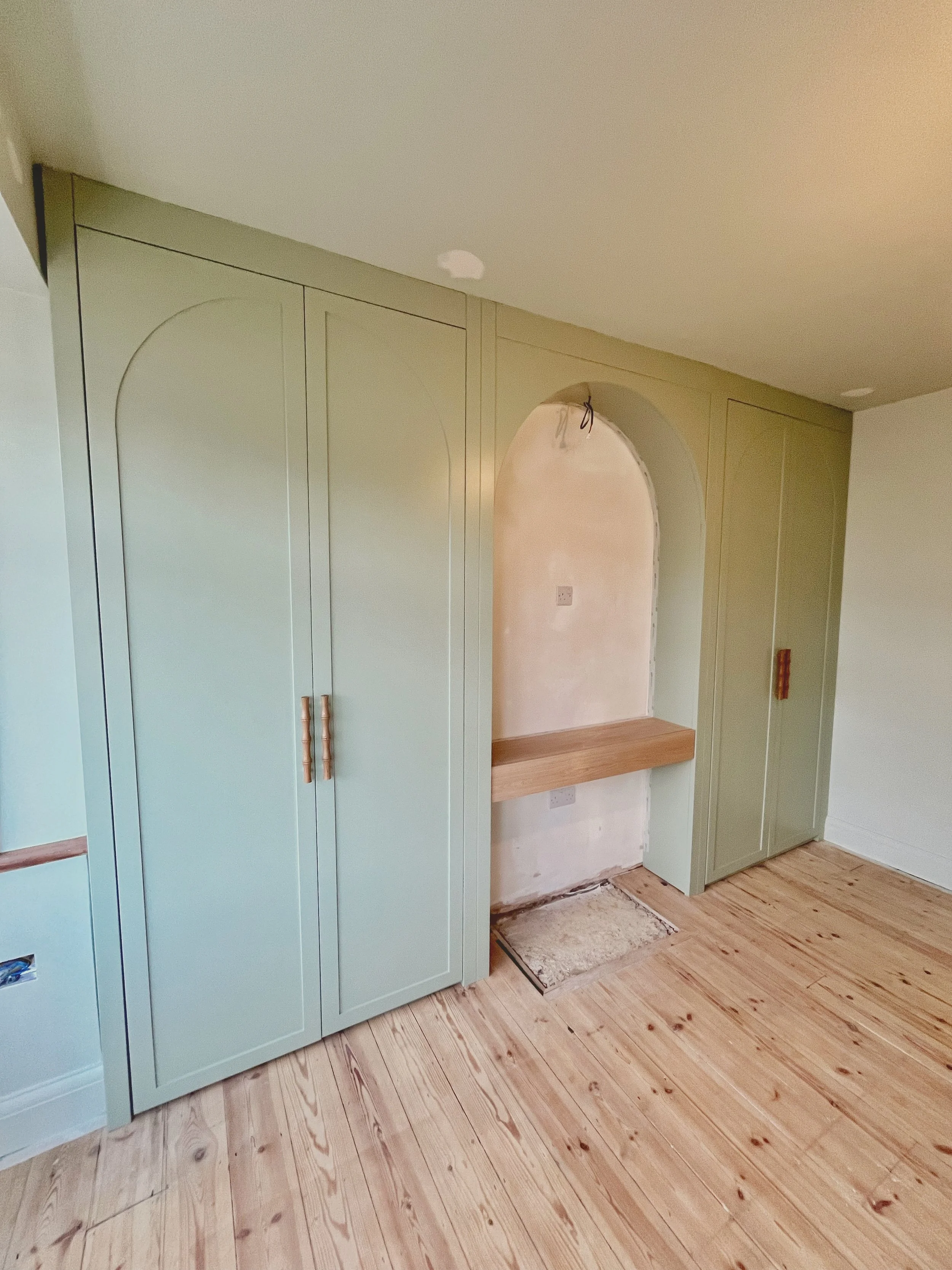 Room with green built-in cabinets and wooden flooring, featuring a small niche with an arched opening and a wooden shelf underneath.