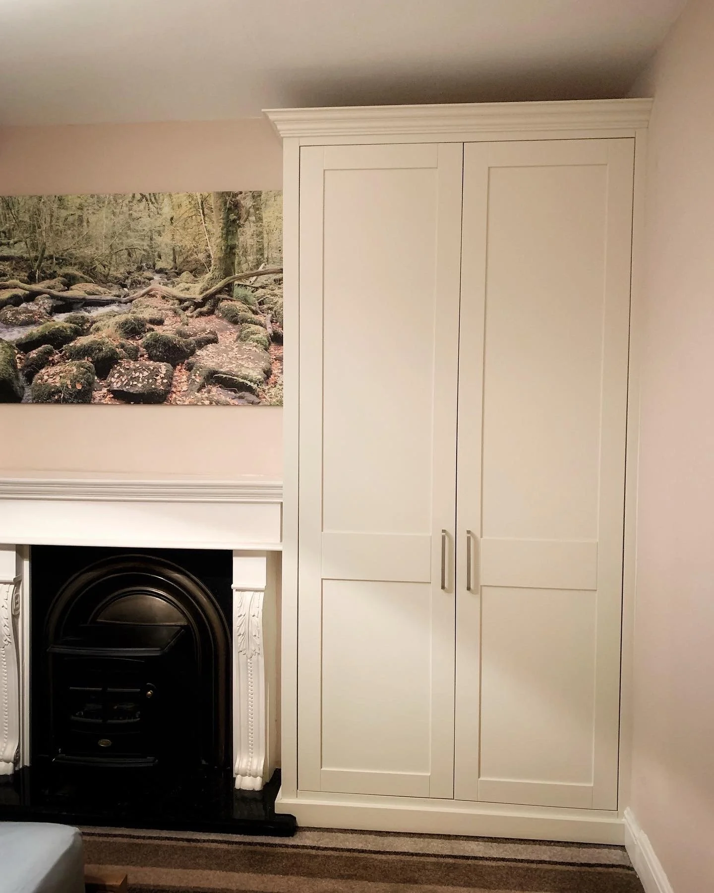 A white built-in wardrobe next to a fireplace with decorative molding in a room with beige walls and a nature landscape poster.
