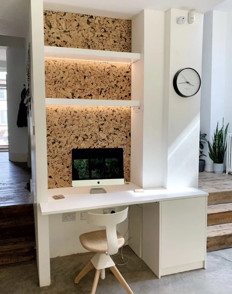 Home office workspace with a white built-in desk, cork wall panels with backlighting, a computer monitor, a white and wood desk chair, a wall clock, and potted plants on wooden steps nearby.