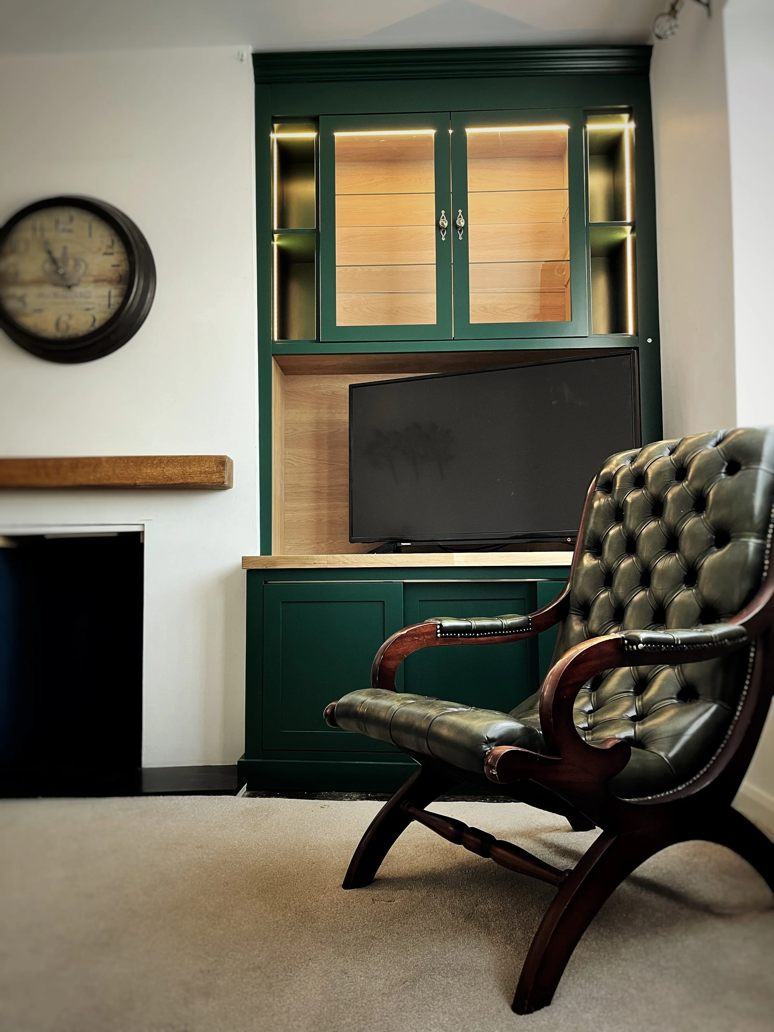 Living room with a dark tufted leather armchair with wooden armrests in front of a green and wood entertainment center, a flat-screen TV, a wall clock, and a beige carpet.
