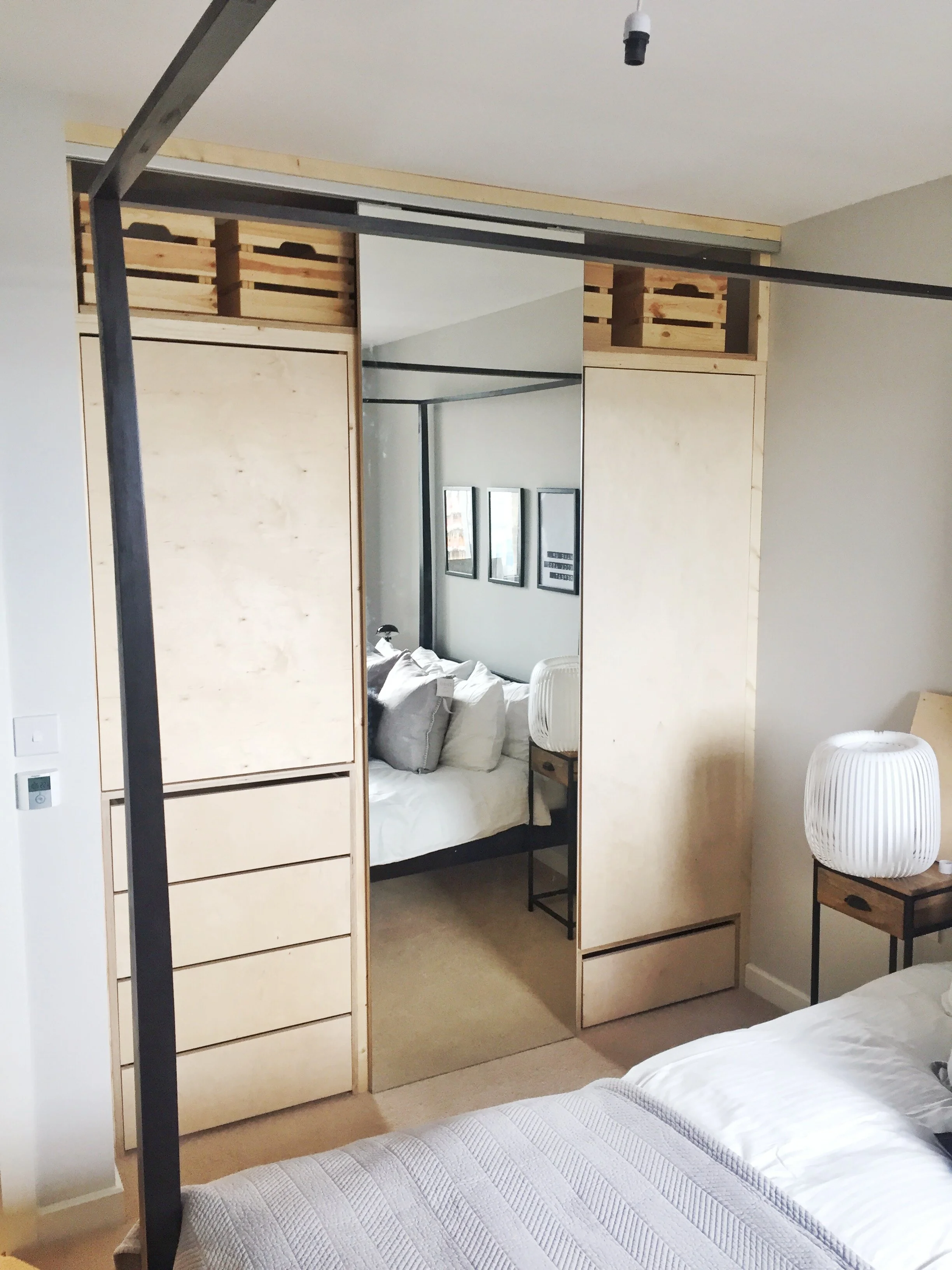 Bedroom with a bed, nightstand, lamp, and a mirrored sliding closet door reflecting the bed and pillows.