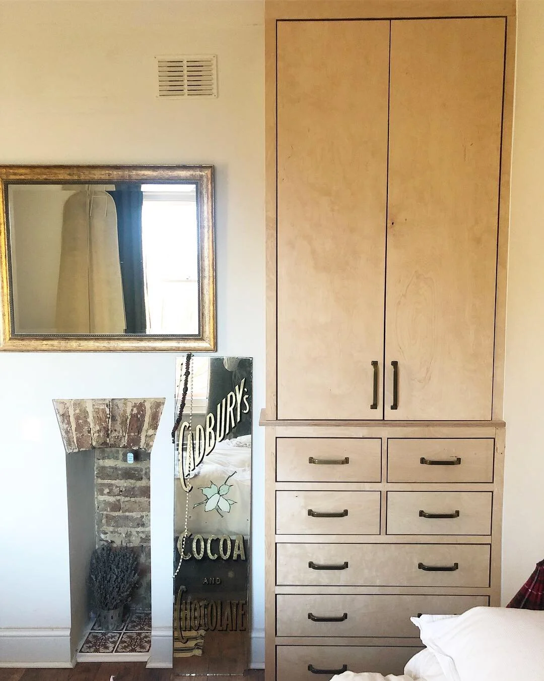 Interior of a room showing a tall wooden cabinet with drawers, a large rectangular mirror with a wooden frame, a decorative mirror with the text "DORIS'S COCOA AND CHOCOLATE," a small window, a wall vent, and part of a bed with white bedding.