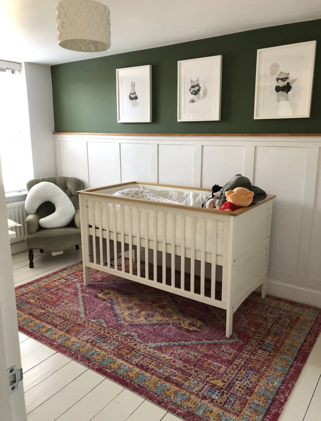 Baby crib in a room with framed animal illustrations on a green wall, a green upholstered armchair with a white neck pillow, on a colorful rug with white wooden floor.