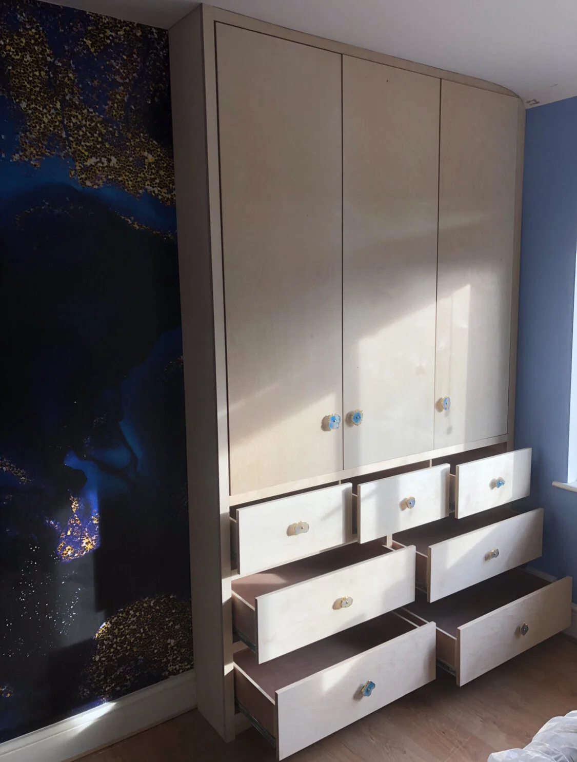 A large light-colored wooden wardrobe with several open drawers, set against a partial wall with a dark blue and black abstract wallpaper design. The wardrobe has multiple doors and the drawers have round handles, some of which are wrapped in blue ta
