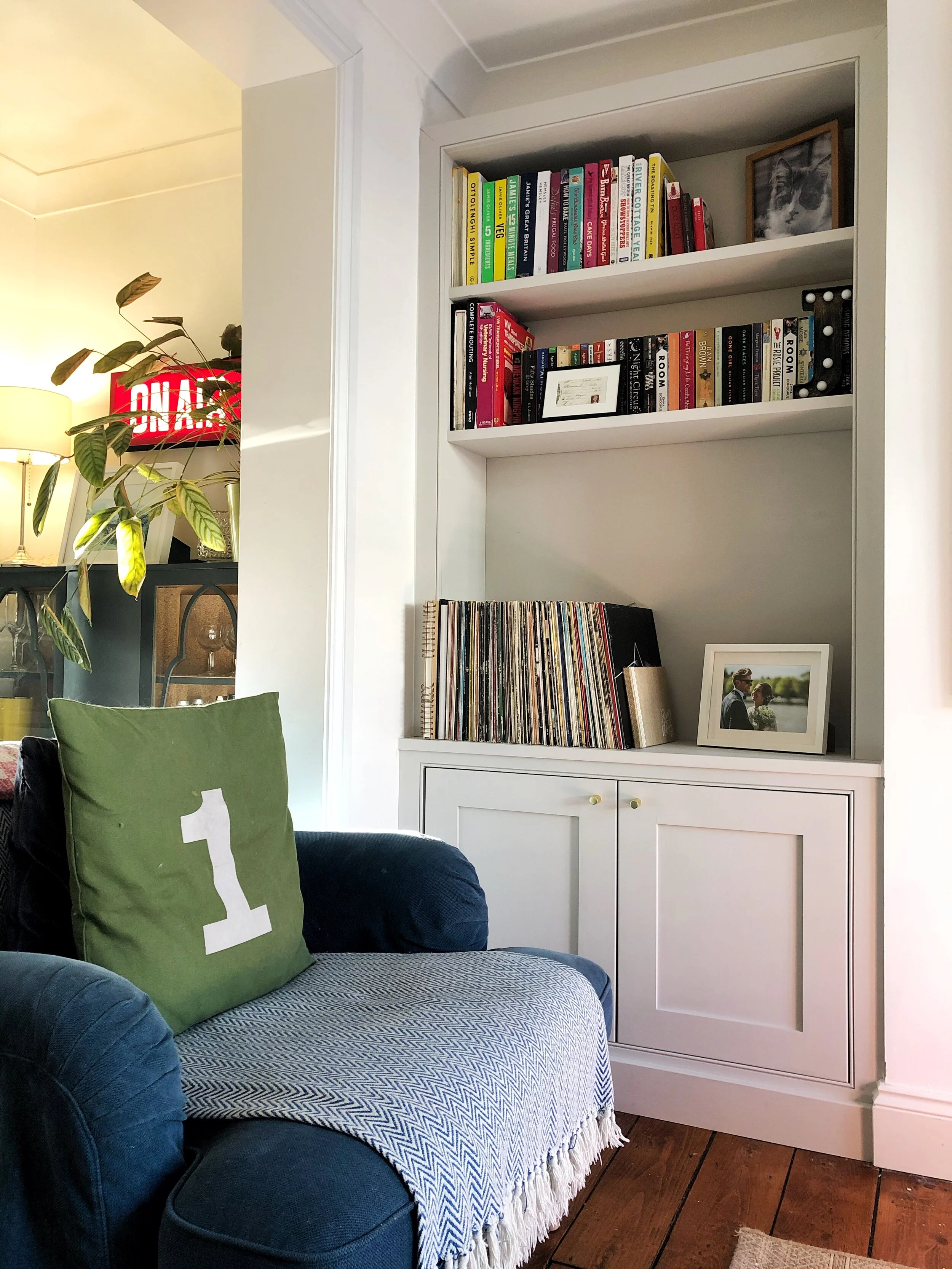 Living room with a navy blue armchair, a green pillow with the number one, a white built-in bookshelf filled with books, a framed photo of a couple, vinyl records, and a part of a plant in the background.