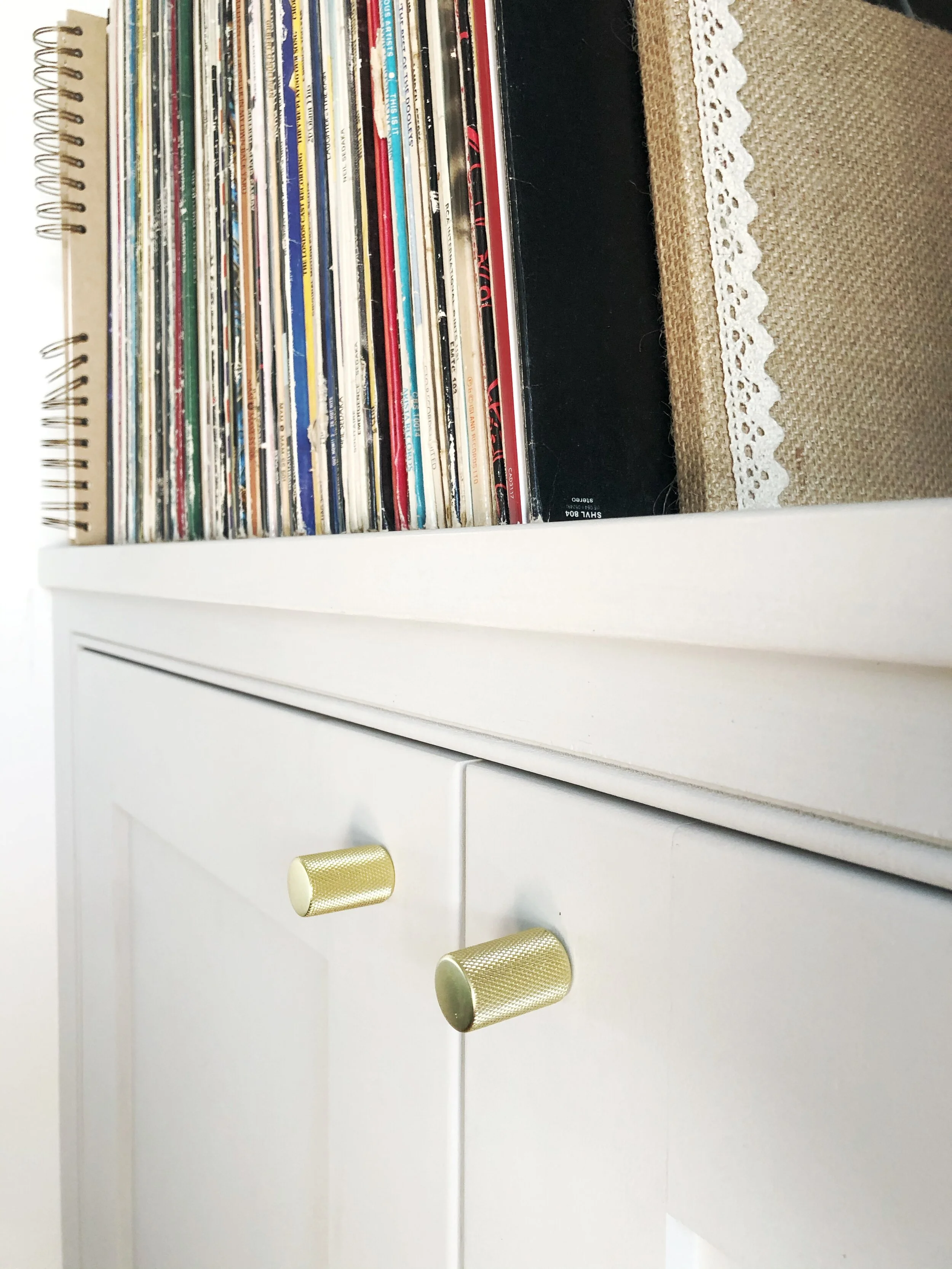 A white cabinet with gold knobs and a top shelf filled with vinyl records and a notepad.