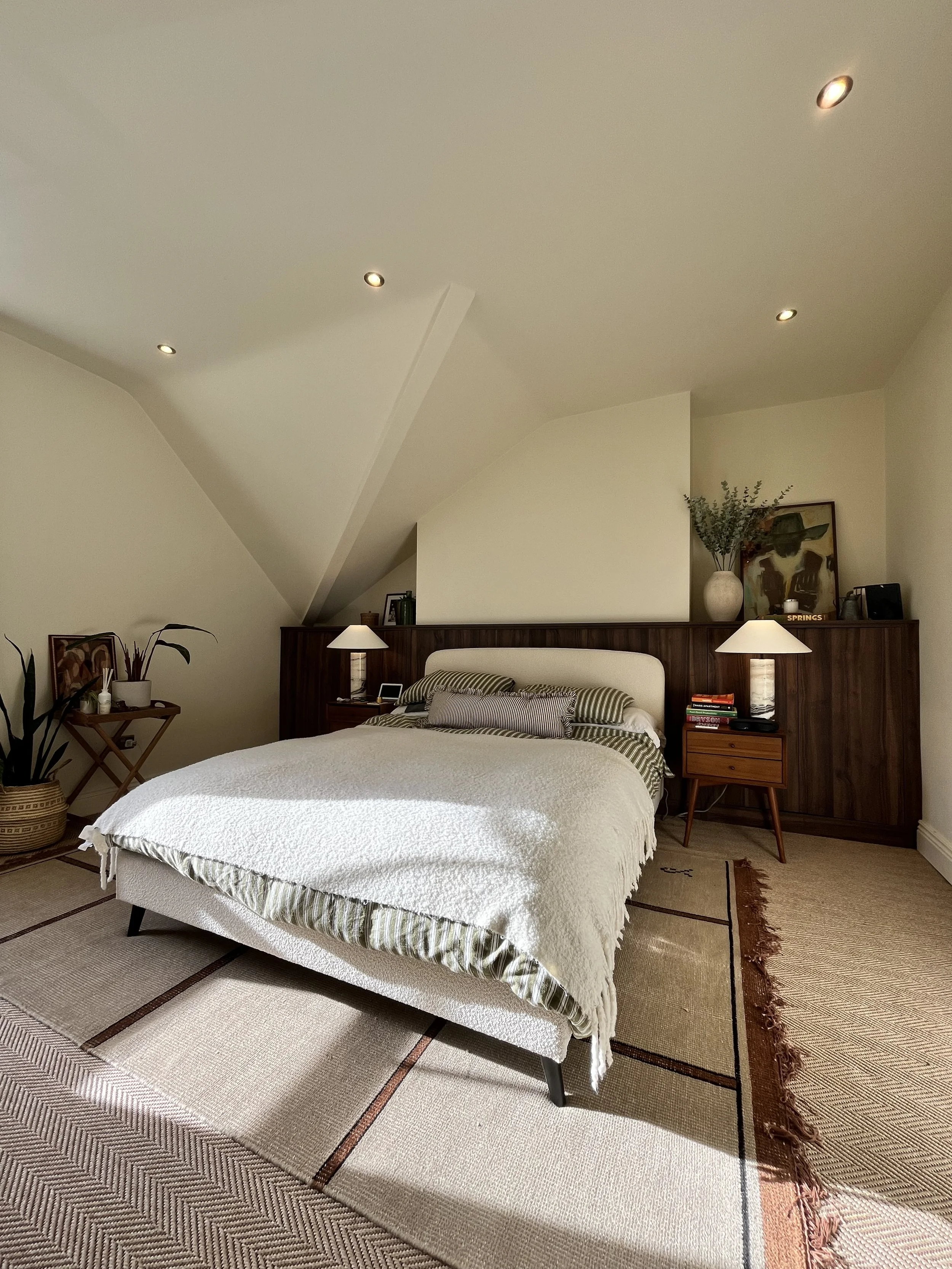 A neatly made bed with striped pillows and a beige blanket in a cozy bedroom with wooden accents, a rug, and minimalistic decor including lamps, artwork, and a plant.