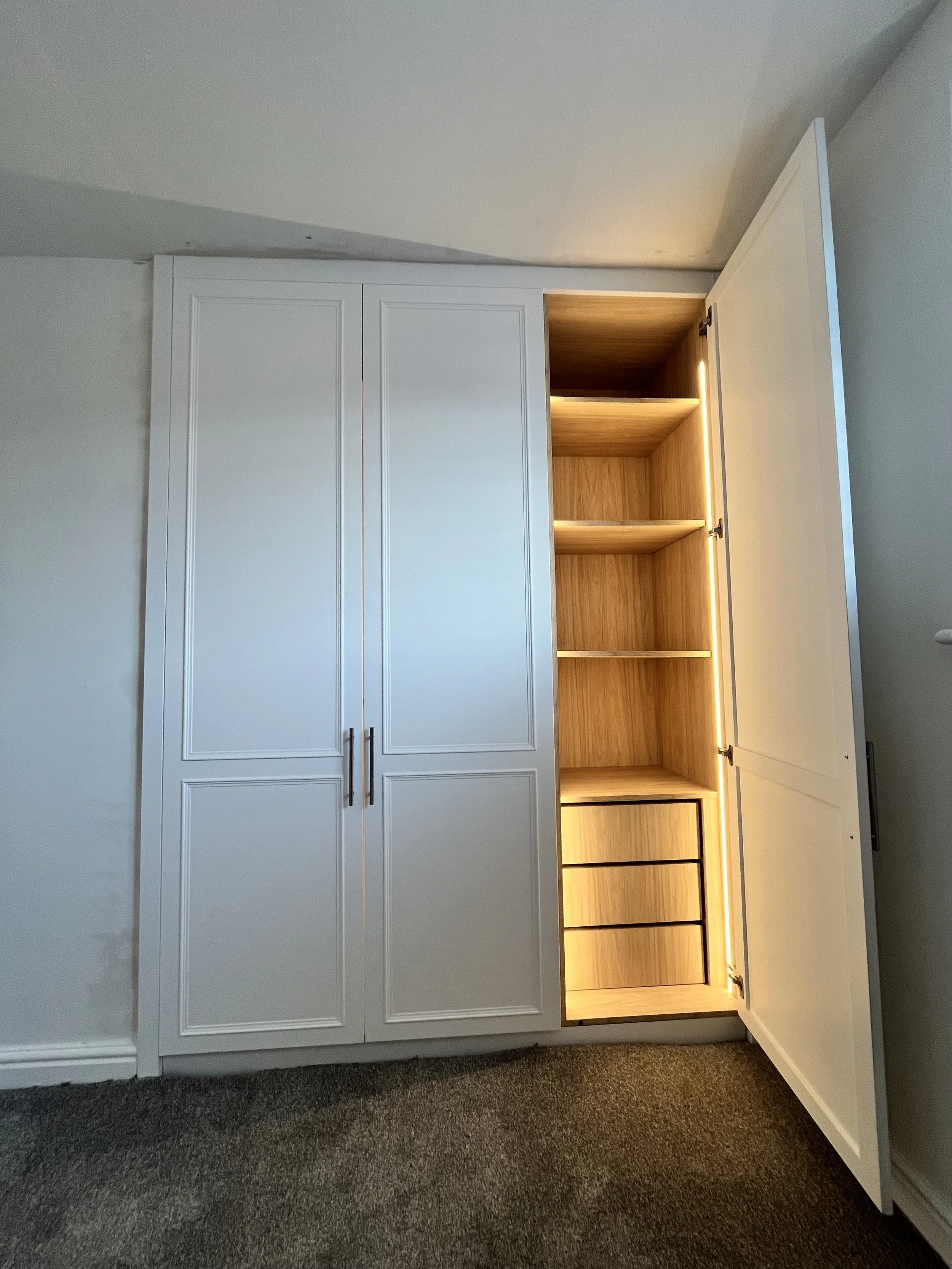 White built-in cabinet with two doors, one open revealing wooden shelves and drawers inside, on a carpeted floor.