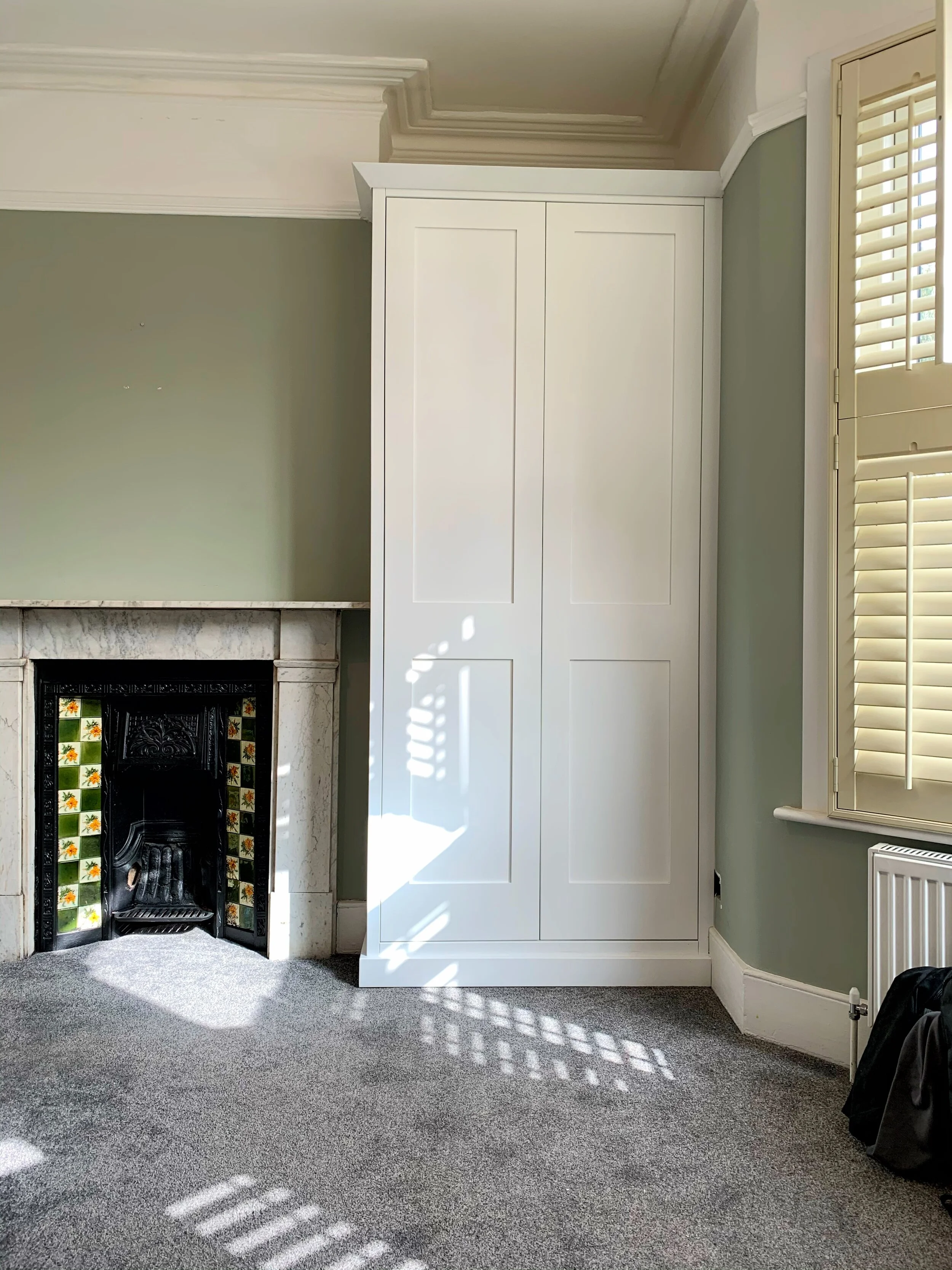 A corner of a room with a fireplace, a tall white cabinet, a window with white shutters, and a gray carpeted floor.